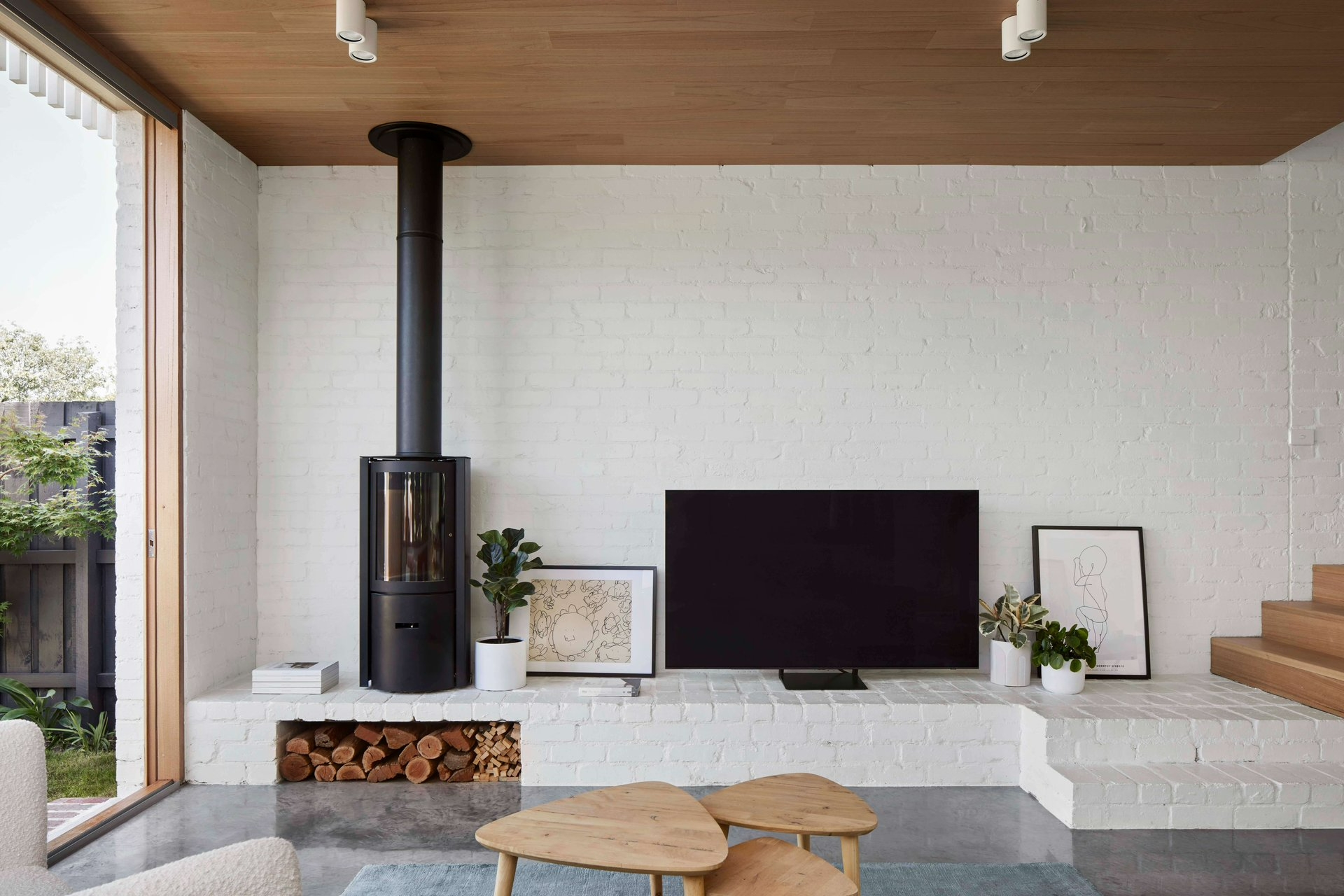 Rosie by Eckersley Architects. Photography by Tess Kelly. Living space with white brick walls, polished concrete flooring, timber coffee tables and black fireplace. 