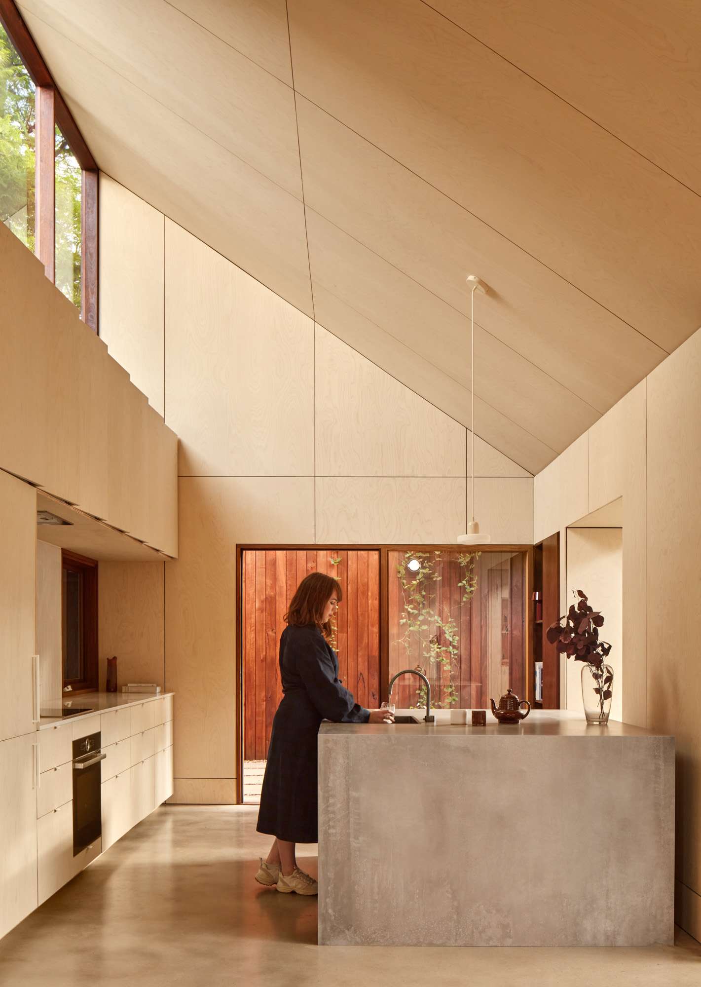 Plywood lined kitchen showing a women filling a glass with water on a concrete island bench.