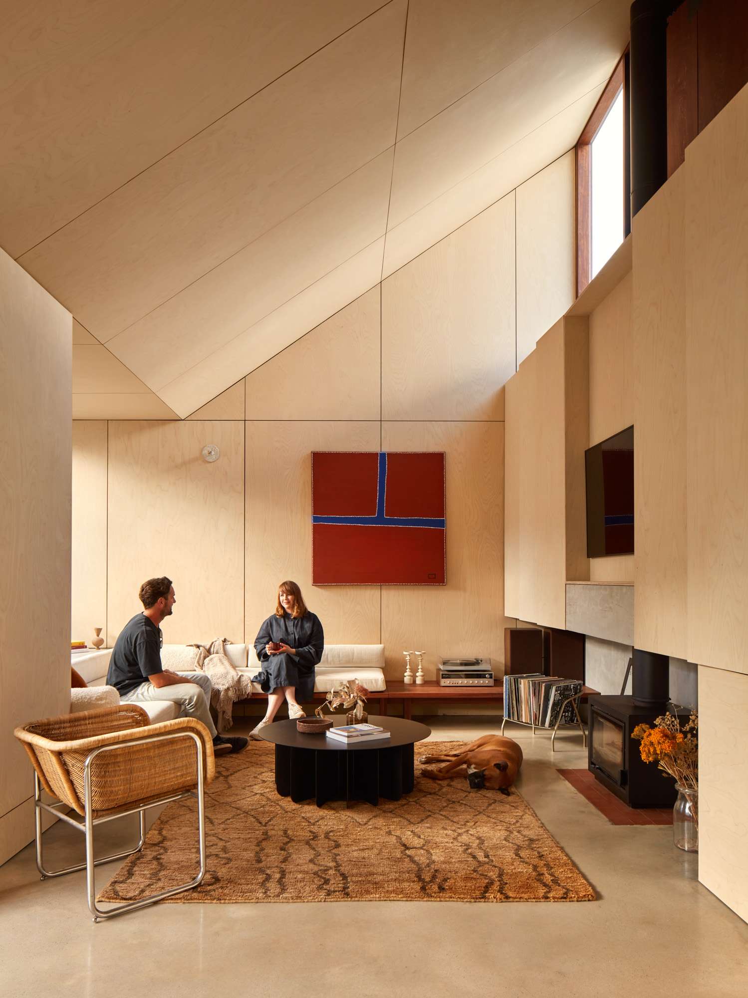 An interior shot of a living space with a high ceiling and a couple sitting on the lounge with a dog on the rug.