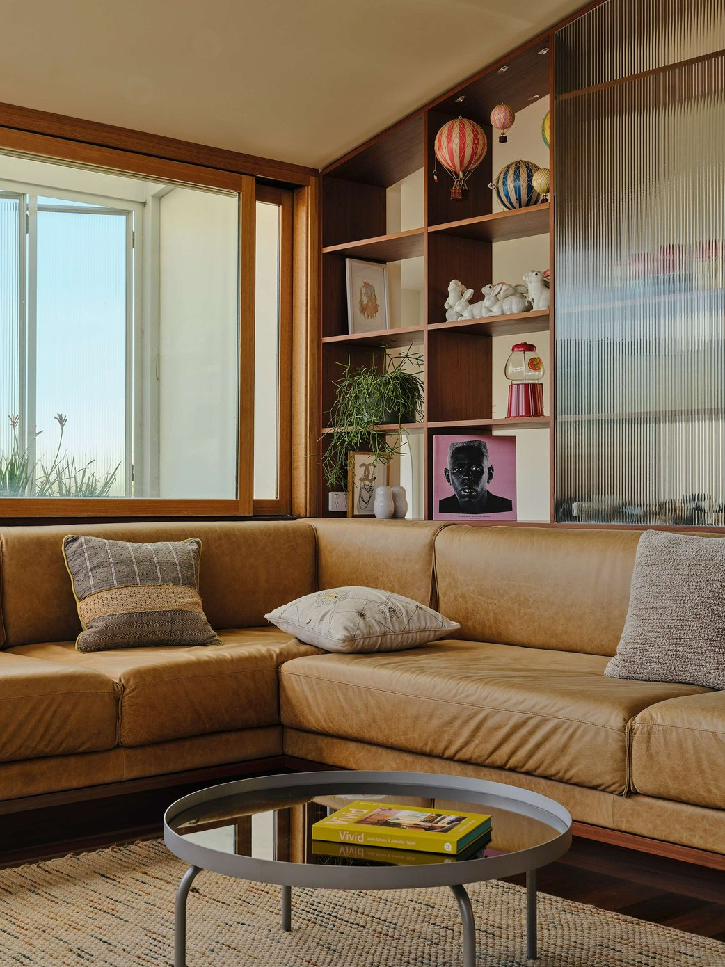 Salmon House by True Story. Photography by Dean Bradley. Living space with tan leather couch, timber cabinetry with fluted glass screen and large timber-framed windows. 