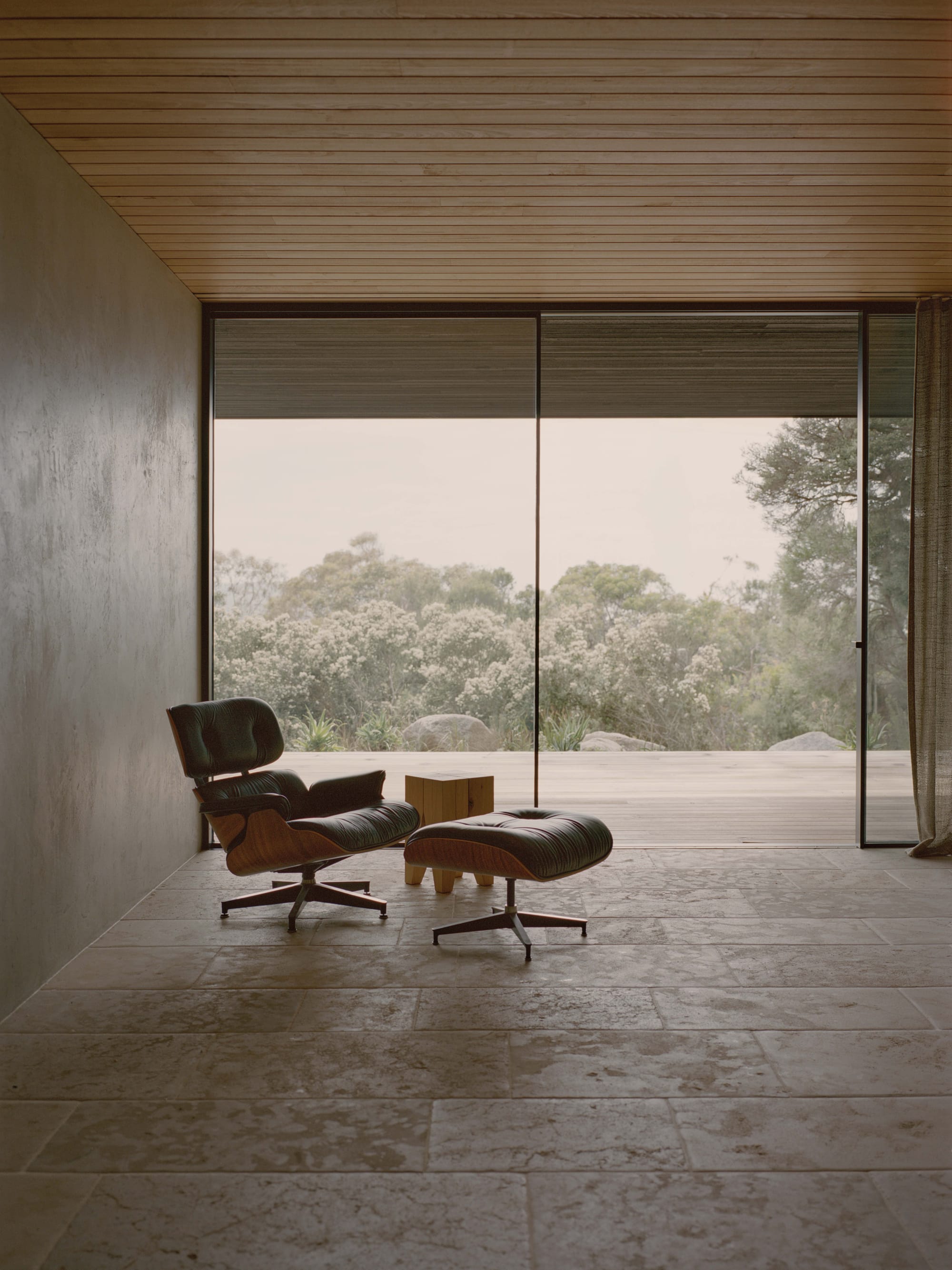 Anglesea House by Eckersley Architects. Photography by Tasha Tylee. Black leather recliner and footstall in space with stone flooring and glass walls, overlooking timber deck and bushland.