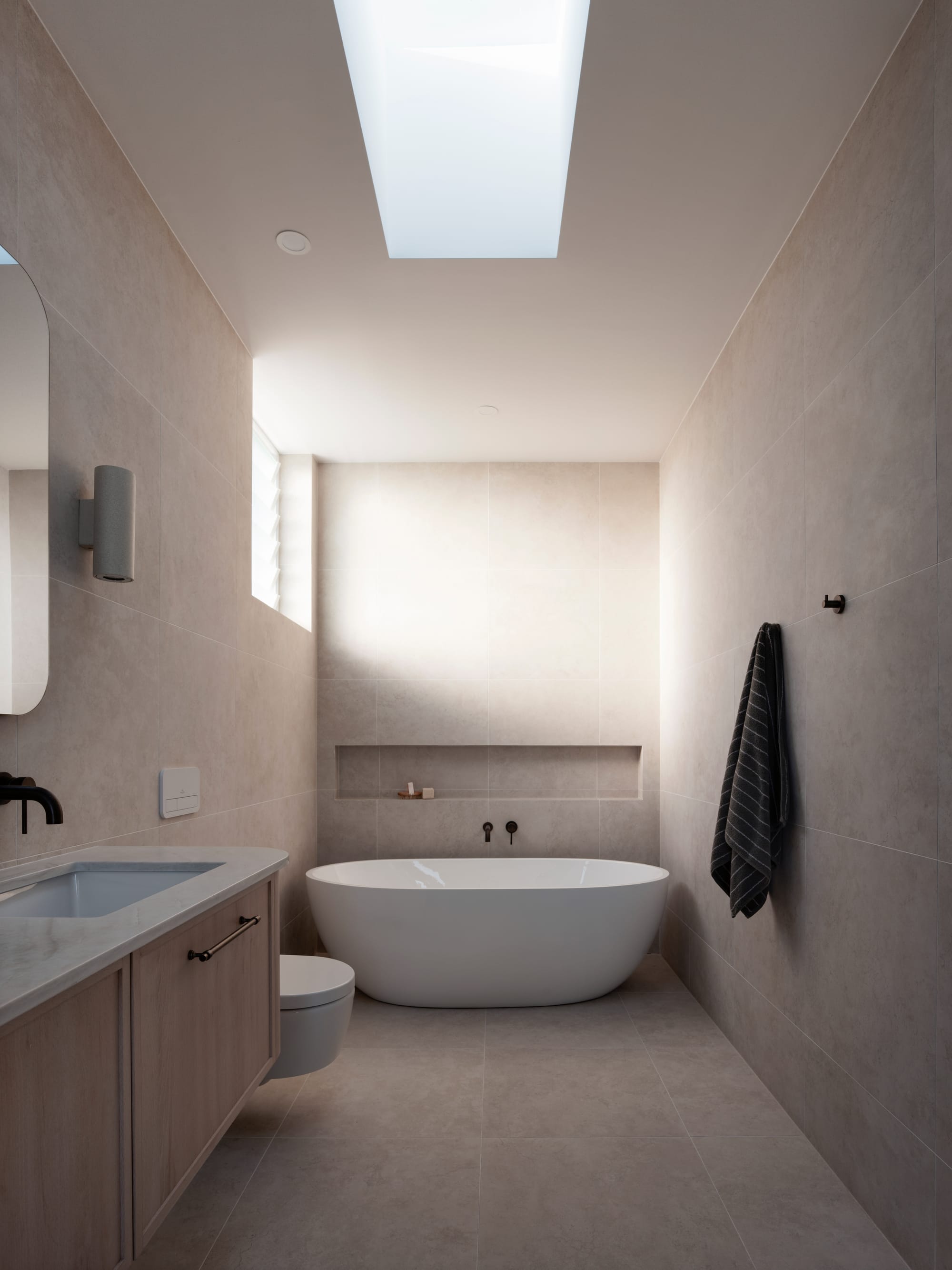 Sienna House by 868 Architects. Photography by Luke Butterly. Bathroom with sandstone tiled walls and floors, freestanding white tub and timber cabinetry. 