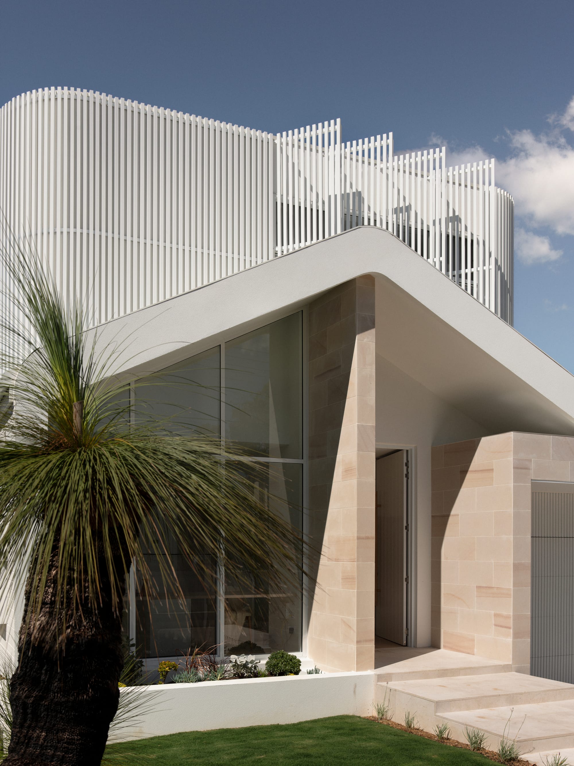 Sienna House by 868 Architects. Photography by Luke Butterly. Front facade of contemporary home with sandstone exterior, white pitched roof, and white timber panelled upper level. 