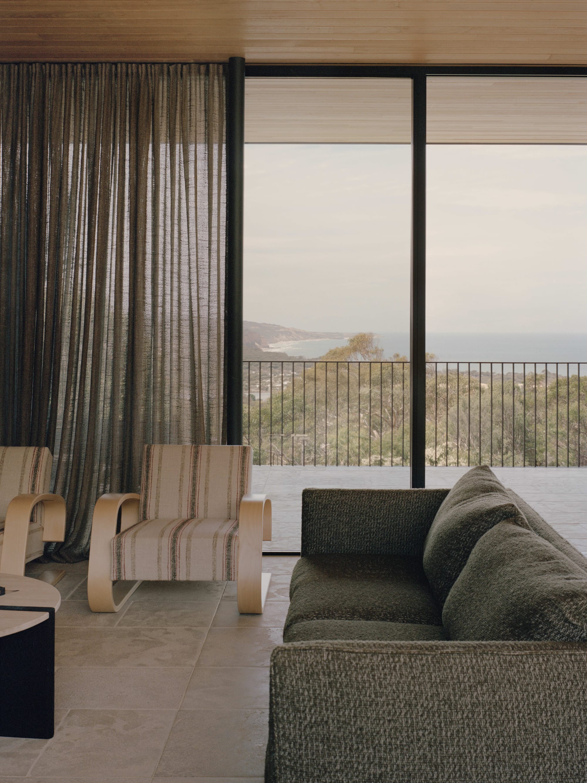 Anglesea House by Eckersley Architects. Photography by Tasha Tylee. Living space with grey couch, timber and striped upholstery armchairs, tiled floors and glass walls.