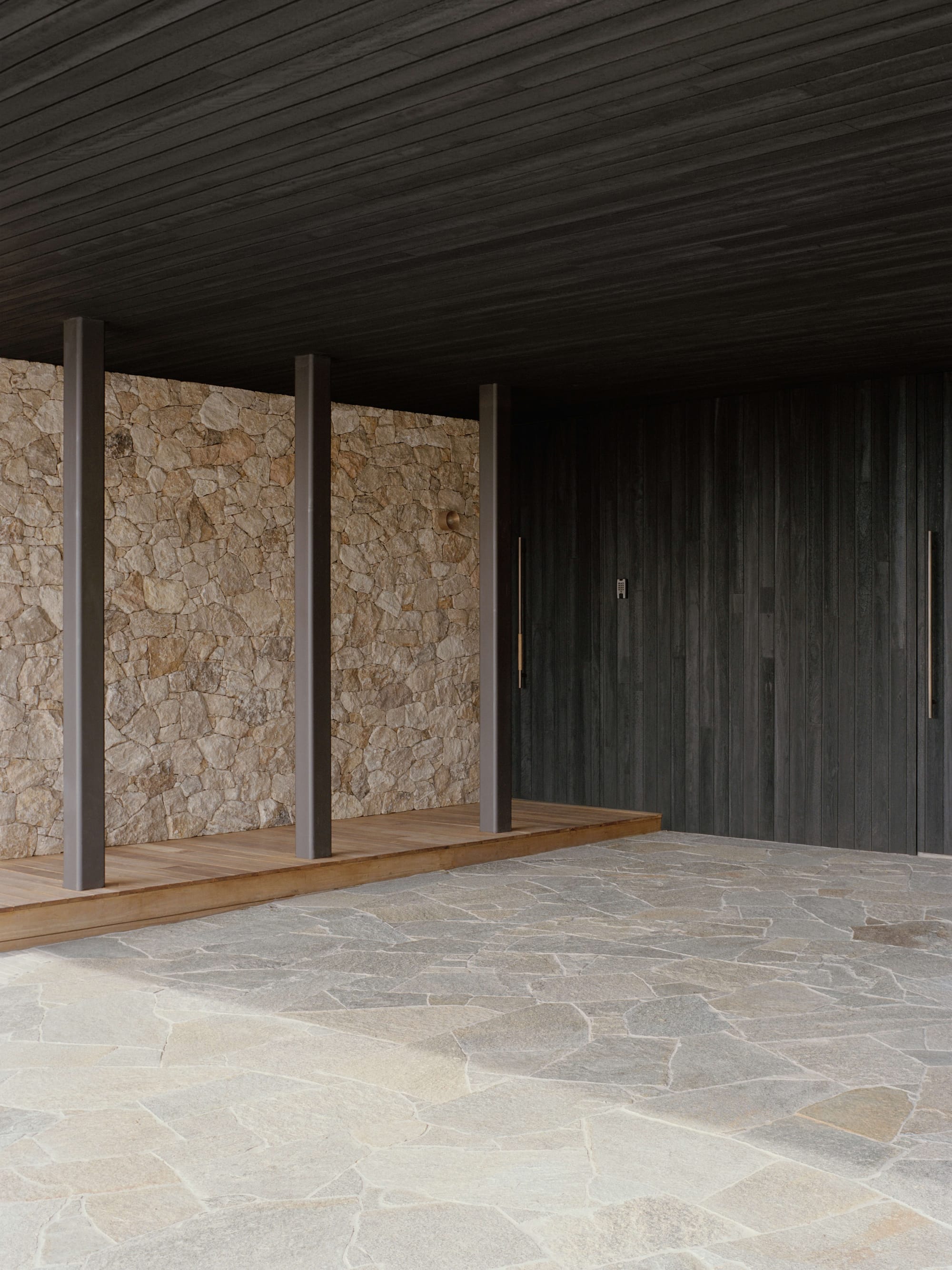 Anglesea House by Eckersley Architects. Photography by Tasha Tylee. Exterior entrance of home with terrazzo flooring, timber decking, stone clad wall, charcoal timber cladding, and minimalistic door handles.