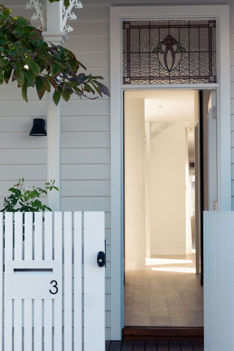 Richmond Heritage Extension by Archive. Photography by Tessa Carroll. Exterior entry of home with grey timber cladding, white timber fence and glass leadlight above open doorway.