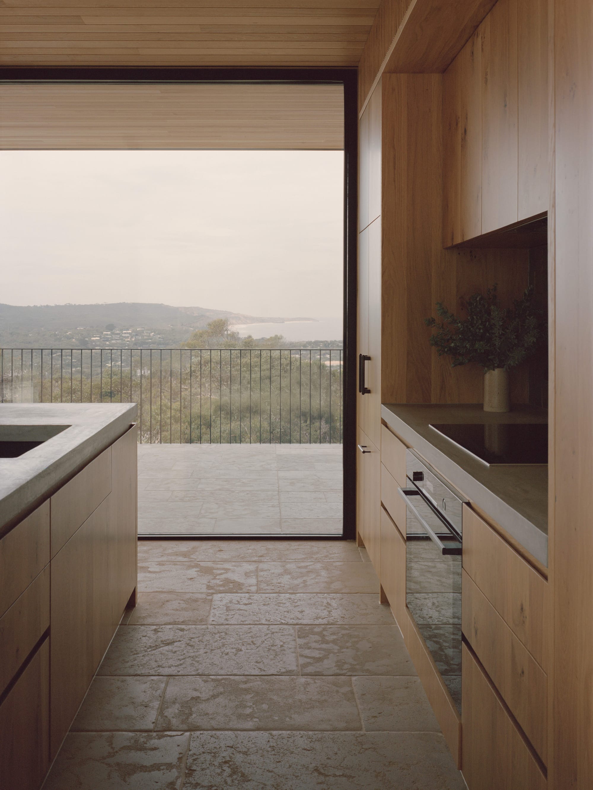 Anglesea House by Eckersley Architects. Photography by Tasha Tylee. Kitchen with stone paver flooring, timber cabinetry, concrete countertops and full-height windows overlooking balcony, with bushland and coastline views.