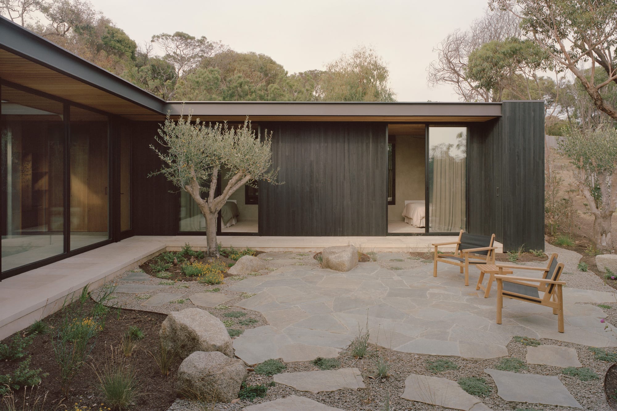 Anglesea House by Eckersley Architects. Photography by Tasha Tylee. Exterior courtyard with stone paver flooring, timber armchairs, and minimal native landscaping, with contemporary home with charcoal timber cladding in background.