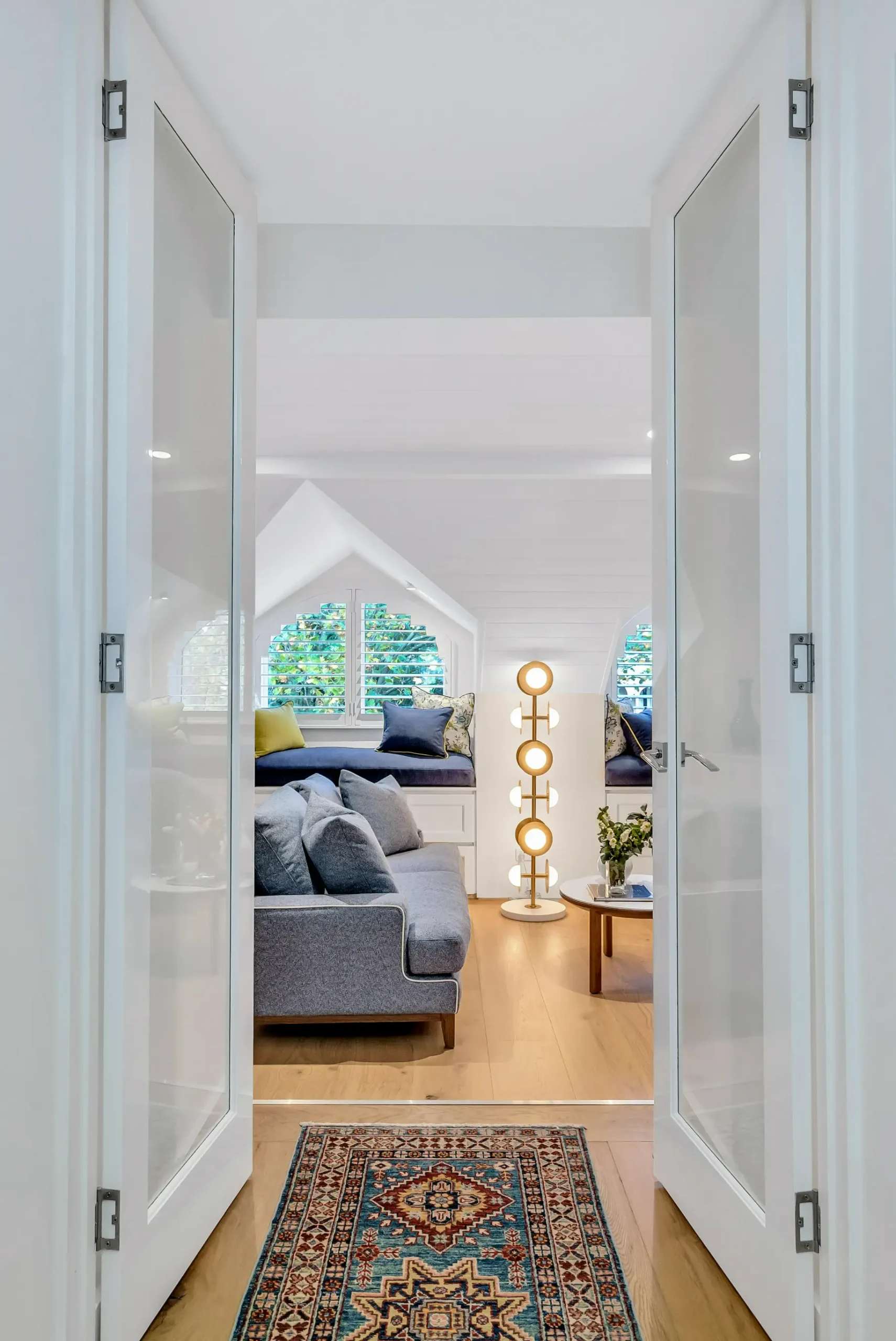 Cozy Elegance by Emotive Designs. View of living room through double white doors, with timber flooring, grey couch, blue bench seating and white ceilings. 