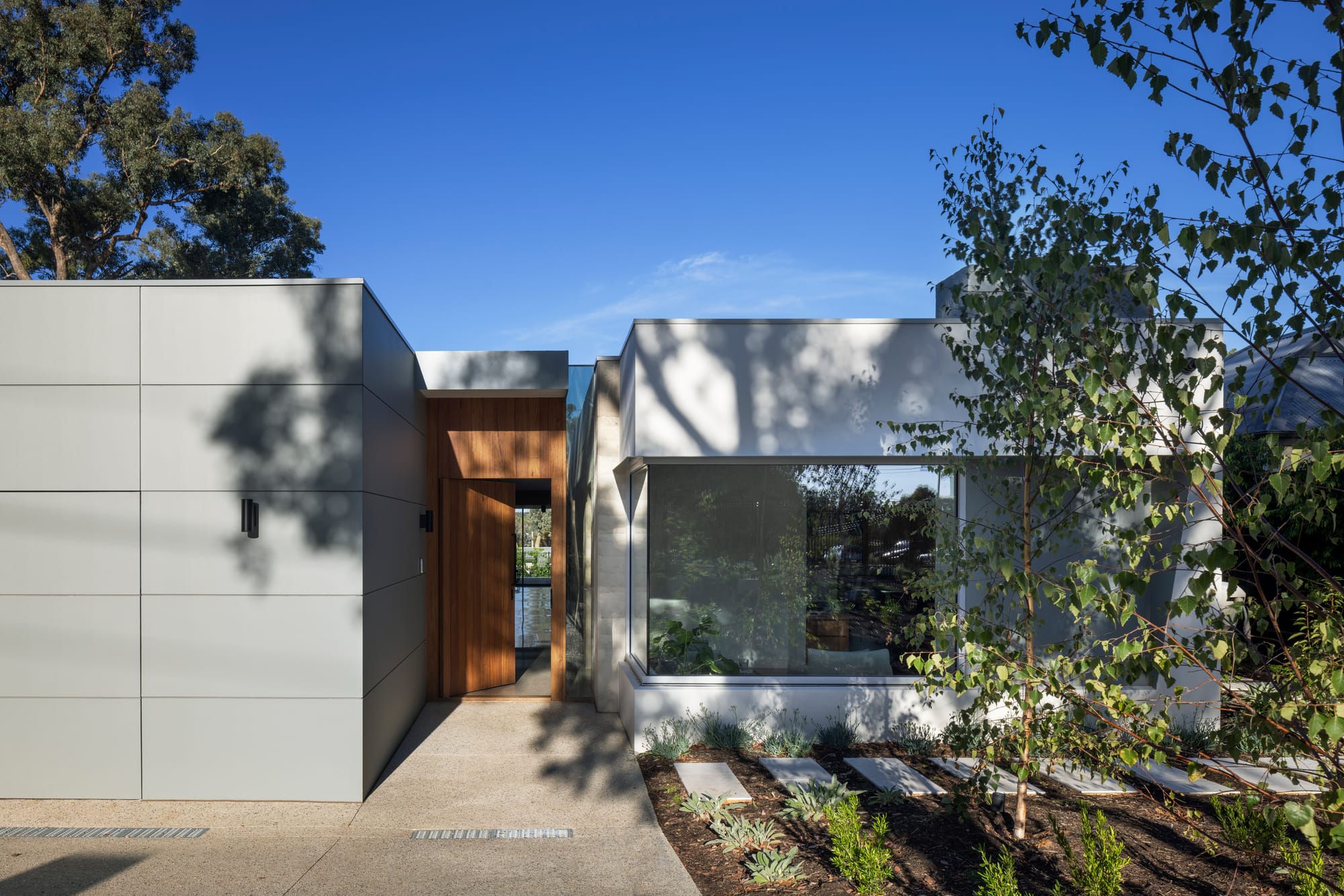 Earth + Sky House by Kirsten Johnstone Architecture. Photography by Tatjana Plitt. Front faacde of single storey home with grey walls, large windows, timber door and pavers through garden. 