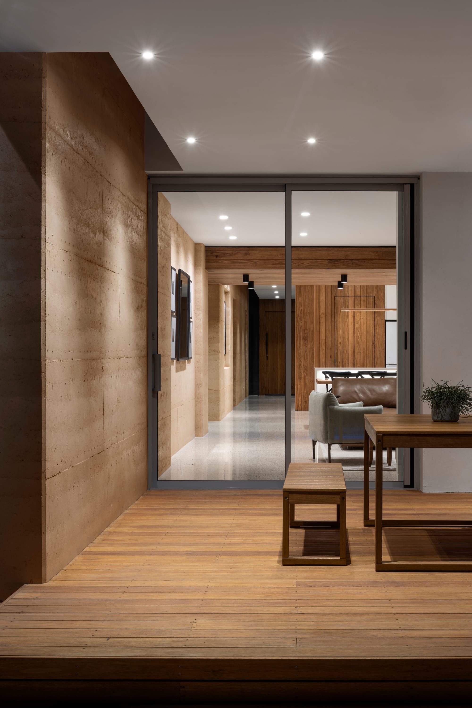 Earth + Sky House by Kirsten Johnstone Architecture. Photography by Tatjana Plitt. Outdoor timber deck at night, with rammed earth walls, timber furniture and light shining out of interior through glass door. 