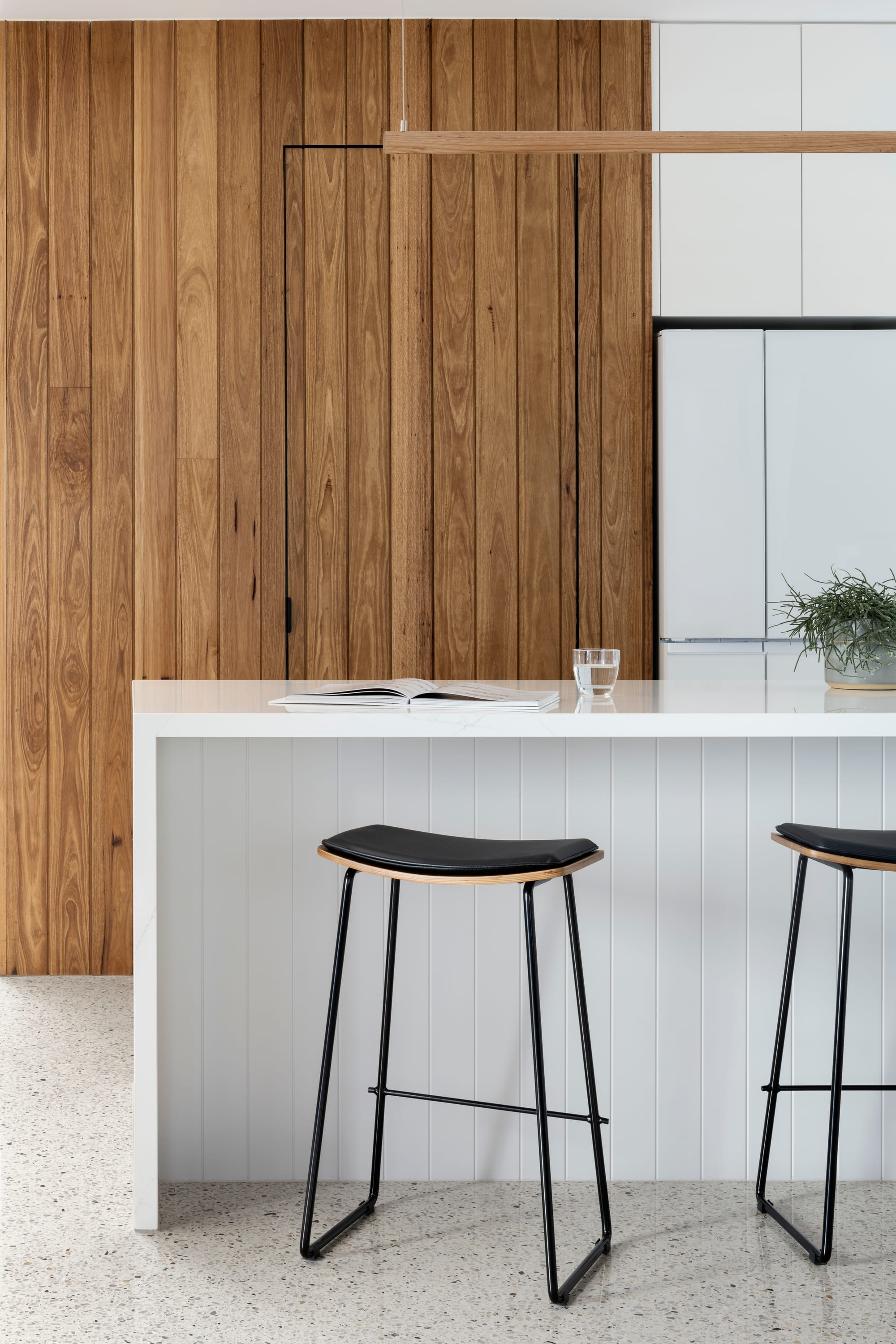 Earth + Sky House by Kirsten Johnstone Architecture. Photography by Tatjana Plitt. Kitchen with polished concerte flooring, timber walls, white cabinetry, and island bench with white timber clad kickboard and stone countertop. 