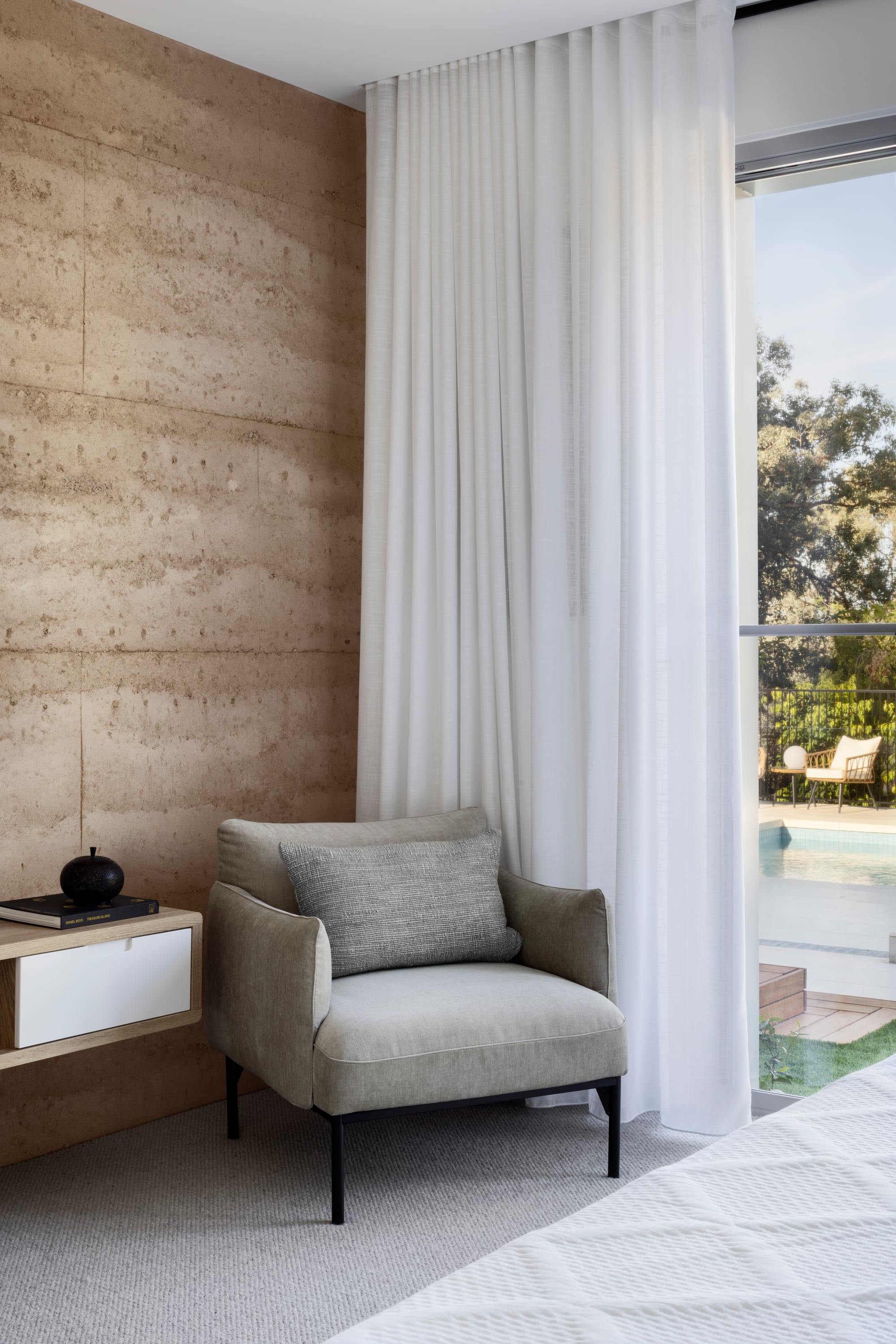 Earth + Sky House by Kirsten Johnstone Architecture. Photography by Tatjana Plitt. Grey armchair in corner of room with white sheer curtains, rammed earth walls, grey carpet and floating timber cabinetry. 