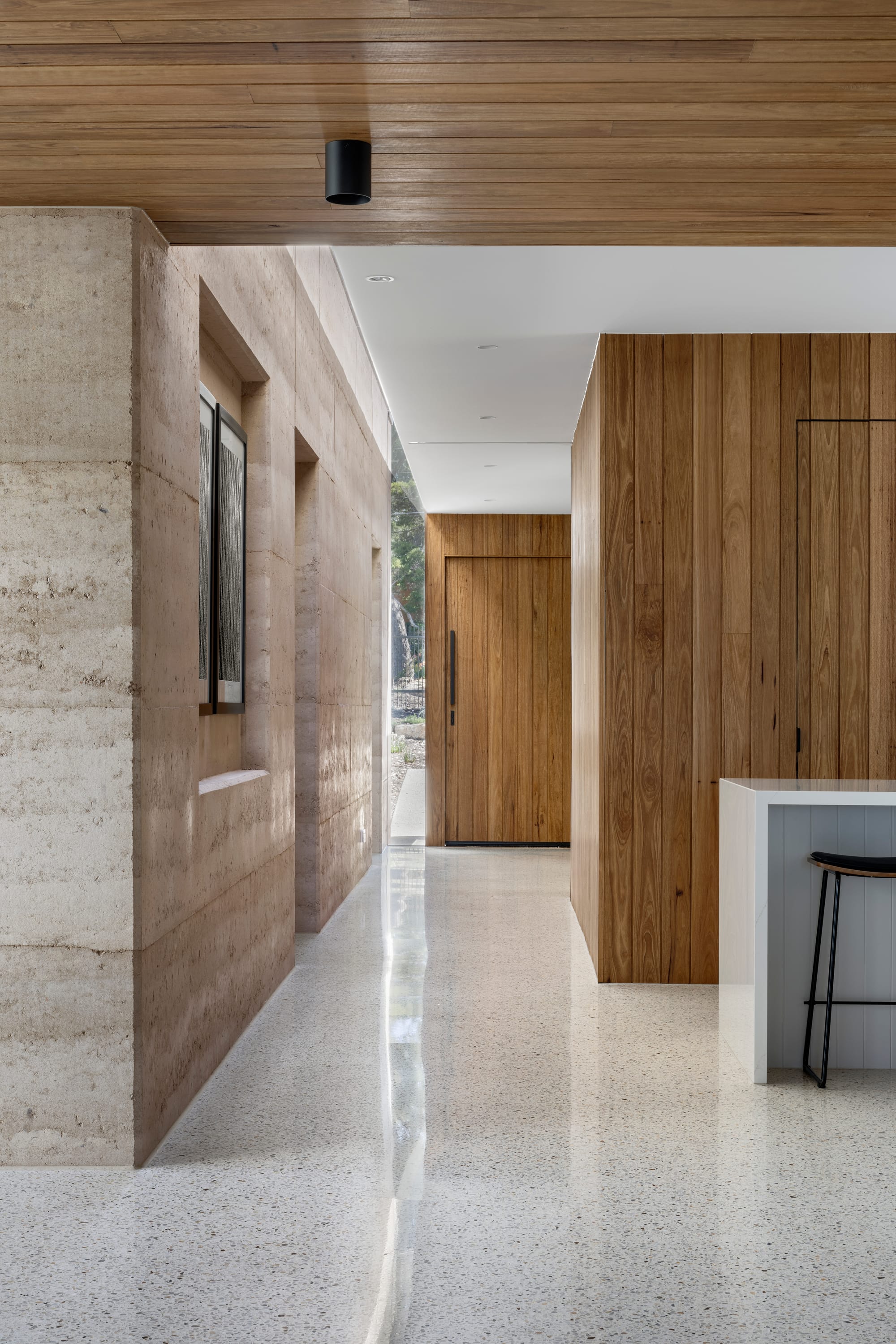 Earth + Sky House by Kirsten Johnstone Architecture. Photography by Tatjana Plitt. Entry and hallway with rammed earth walls to left, timber clad walls to right, polished concrete throughout, and timber front door. 
