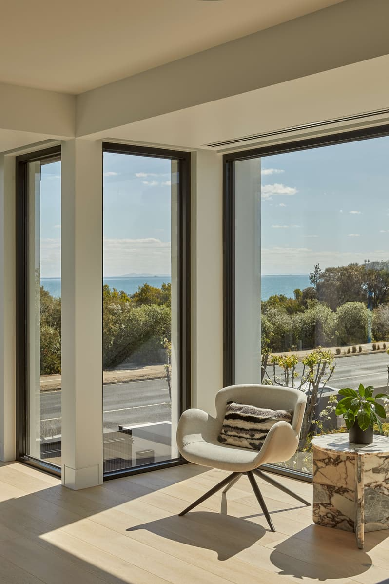 Bay View House by Samuel Architecture. Photography by Dave Kulesza. White armchair with stone side table in front of full height windows overlooking road, bushland and ocean. 