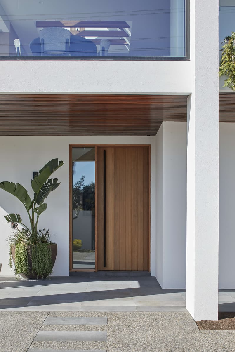 Bay View House by Samuel Architecture. Photography by Dave Kulesza. Exterior entrance of home with timber door, black handle, large potted plant and white walls. 