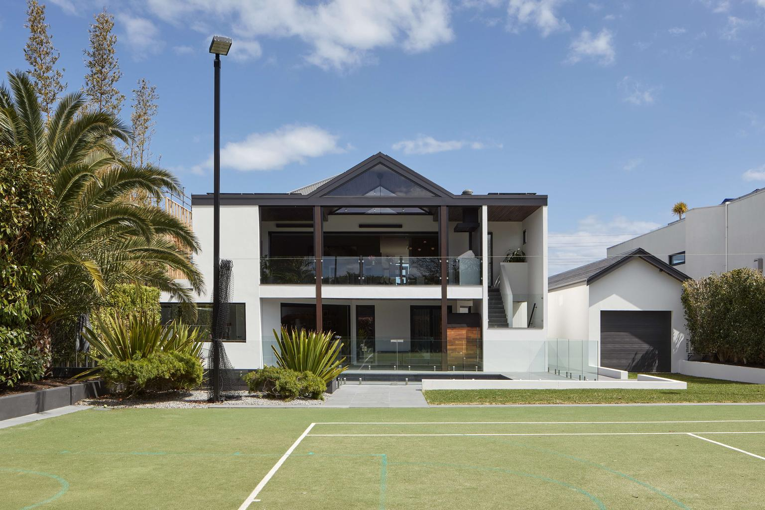 Bay View House by Samuel Architecture. Photography by Dave Kulesza. Exterior facade of double storey modern home with black roof, with grassed tennis court in foreground. 
