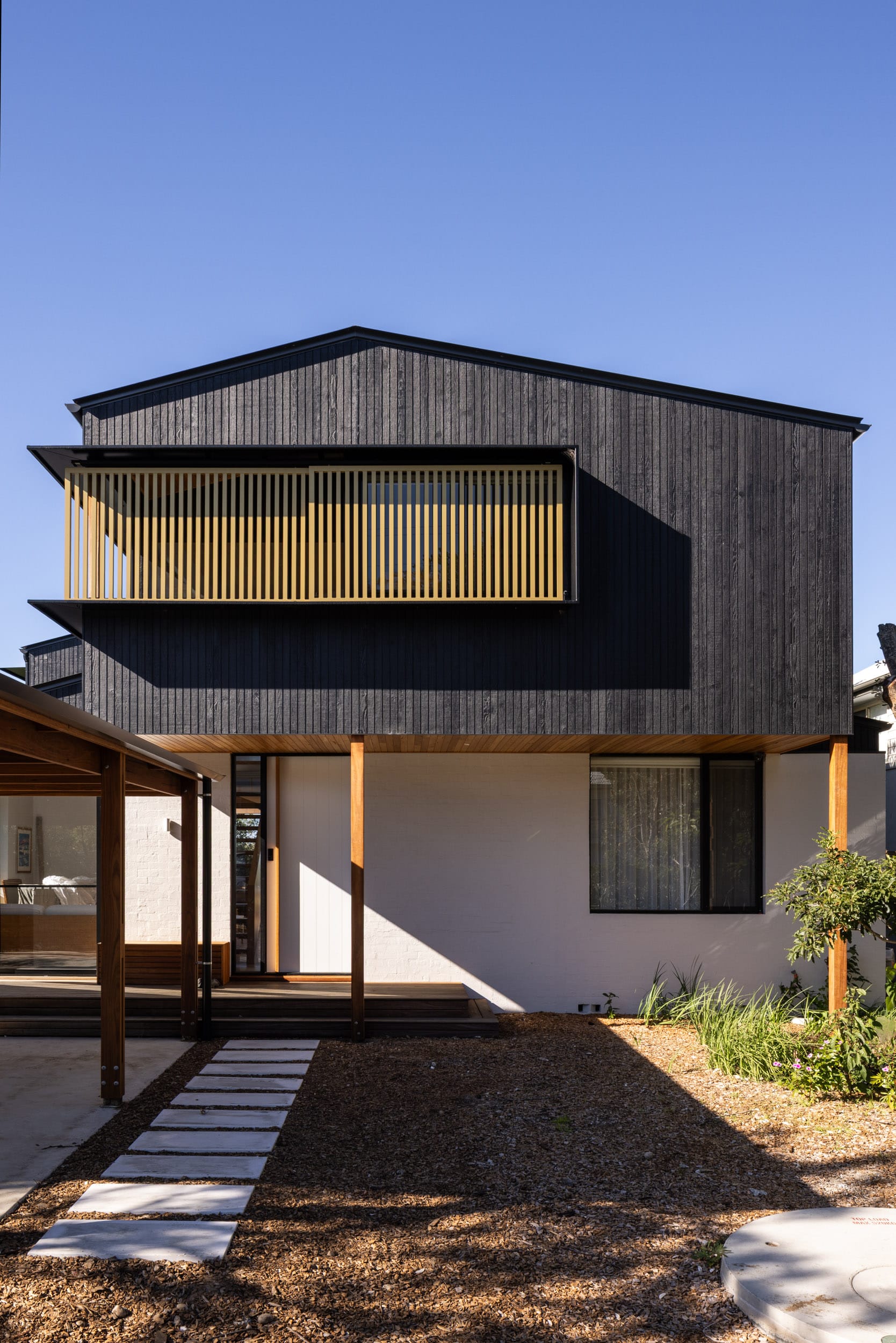 Bawley Point by Watershed Architects. Photography by Simon Whitbread. Double storey home with white brick lower-level facade, charcoal timber clad upper level, and grey stone pavers through mulch to timber deck.   