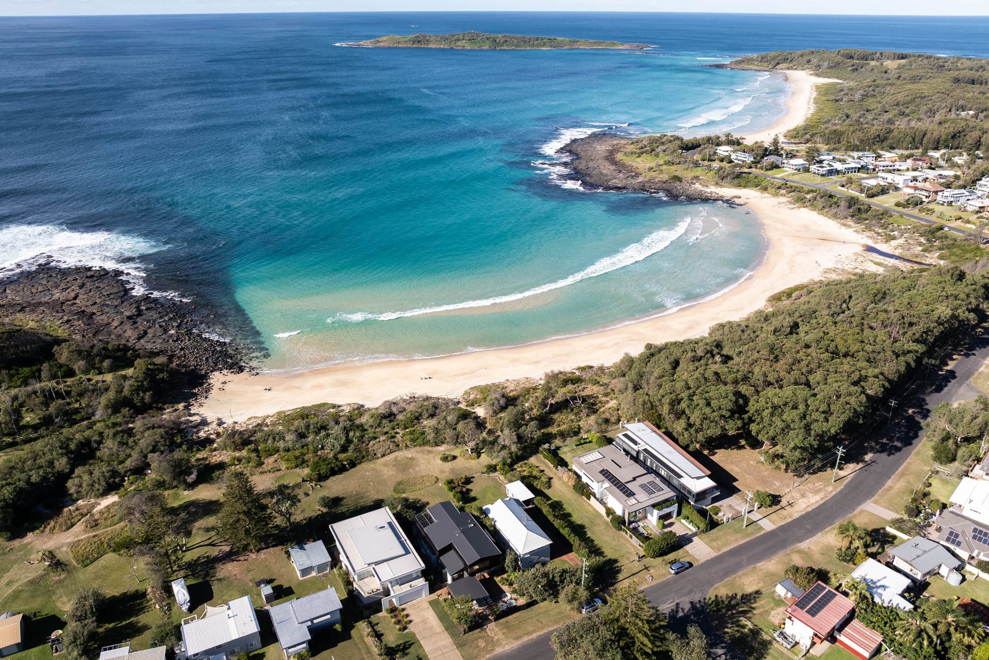 Bawley Point by Watershed Architects. Photography by Simon Whitbread. Aerial view of coastline and beach, with houses bordering onto grass running alongside beach. 
