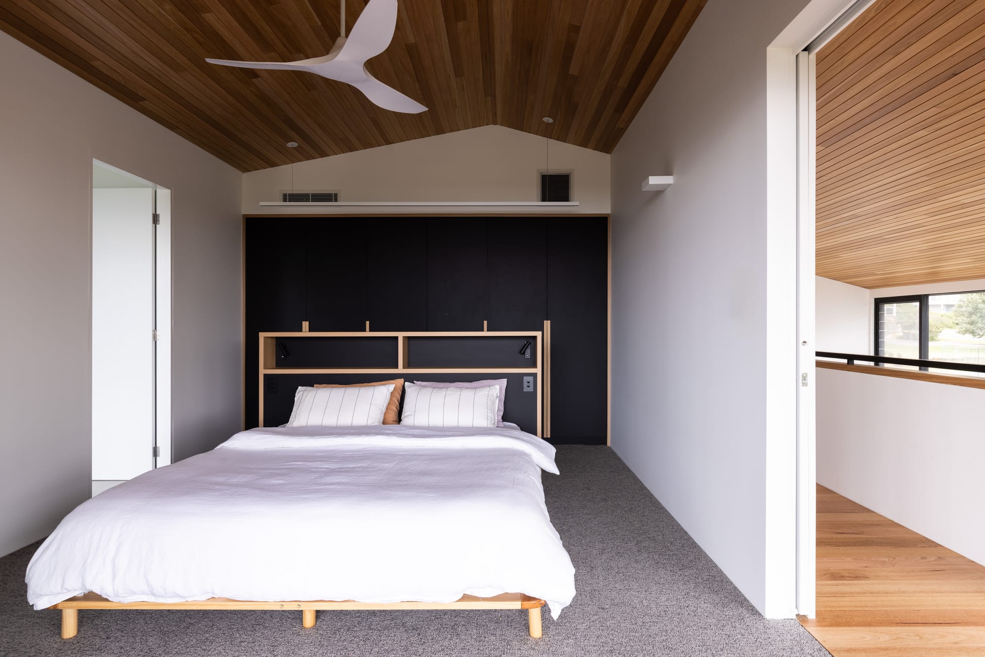 Bawley Point by Watershed Architects. Photography by Simon Whitbread. Bedroom with grey carpets, black integrated cabinetry and bedhead, timber bed with white linen, timber clad ceiling and white walls.  