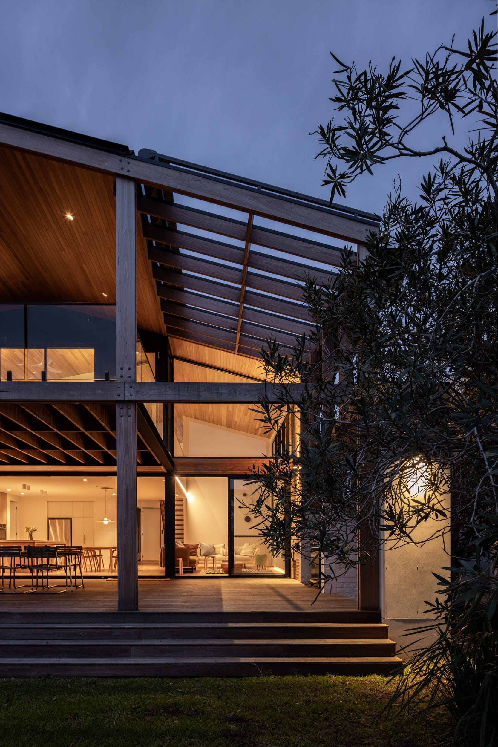 Bawley Point by Watershed Architects. Photography by Simon Whitbread. Rear facade of contemporary double-storey home at night, with warm light shining from interior through large glass windows across eternal timber deck. 
