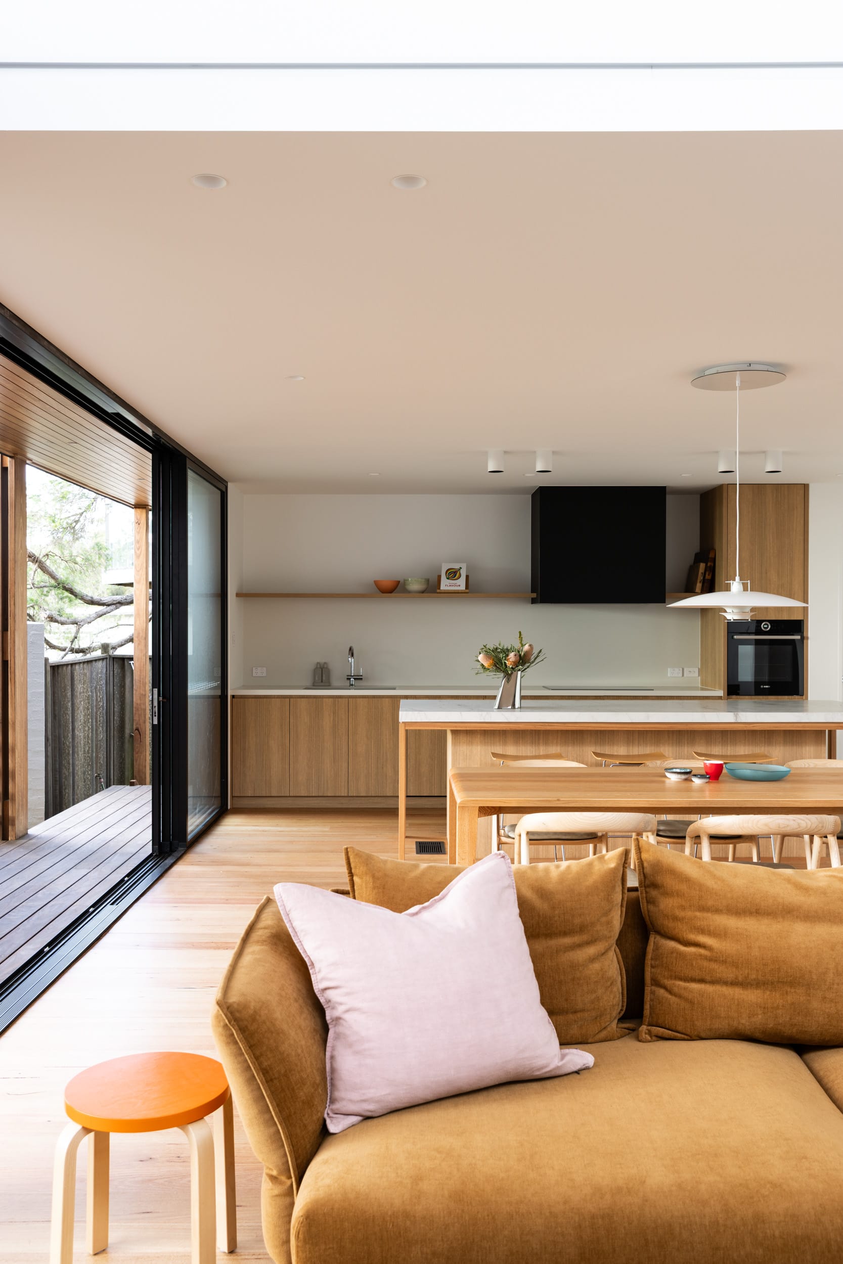 Bawley Point by Watershed Architects. Photography by Simon Whitbread. Mustard yellow suede couch with white cushion in foreground, with kitchen and dining room in the background, with timber cabinetry and flooring throughout. 