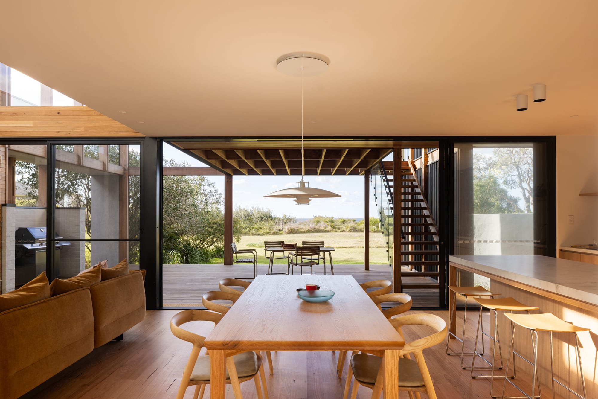 Bawley Point by Watershed Architects. Photography by Simon Whitbread. Timber dining table in centre of open plan kitchen, dining and living area, opening onto timber deck through large sliding glass doors. 
