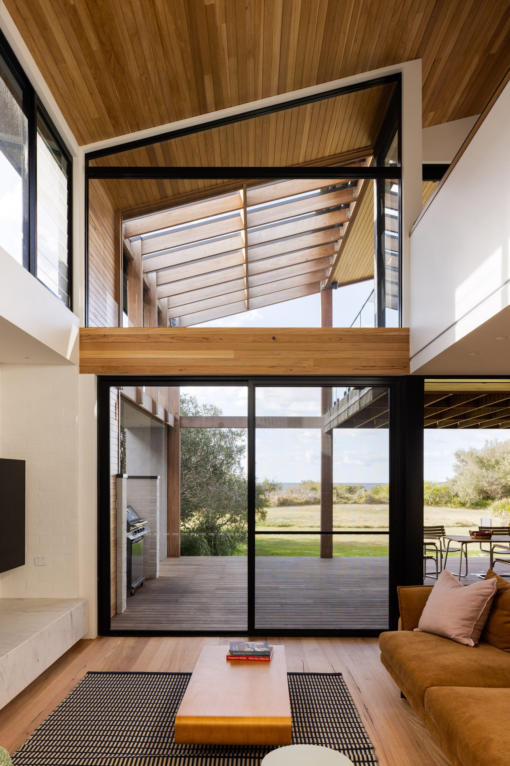 Bawley Point by Watershed Architects. Photography by Simon Whitbread. Living space with timber flooring, tan suede couch and double height glass windows overlooking timber deck and native bushland. 