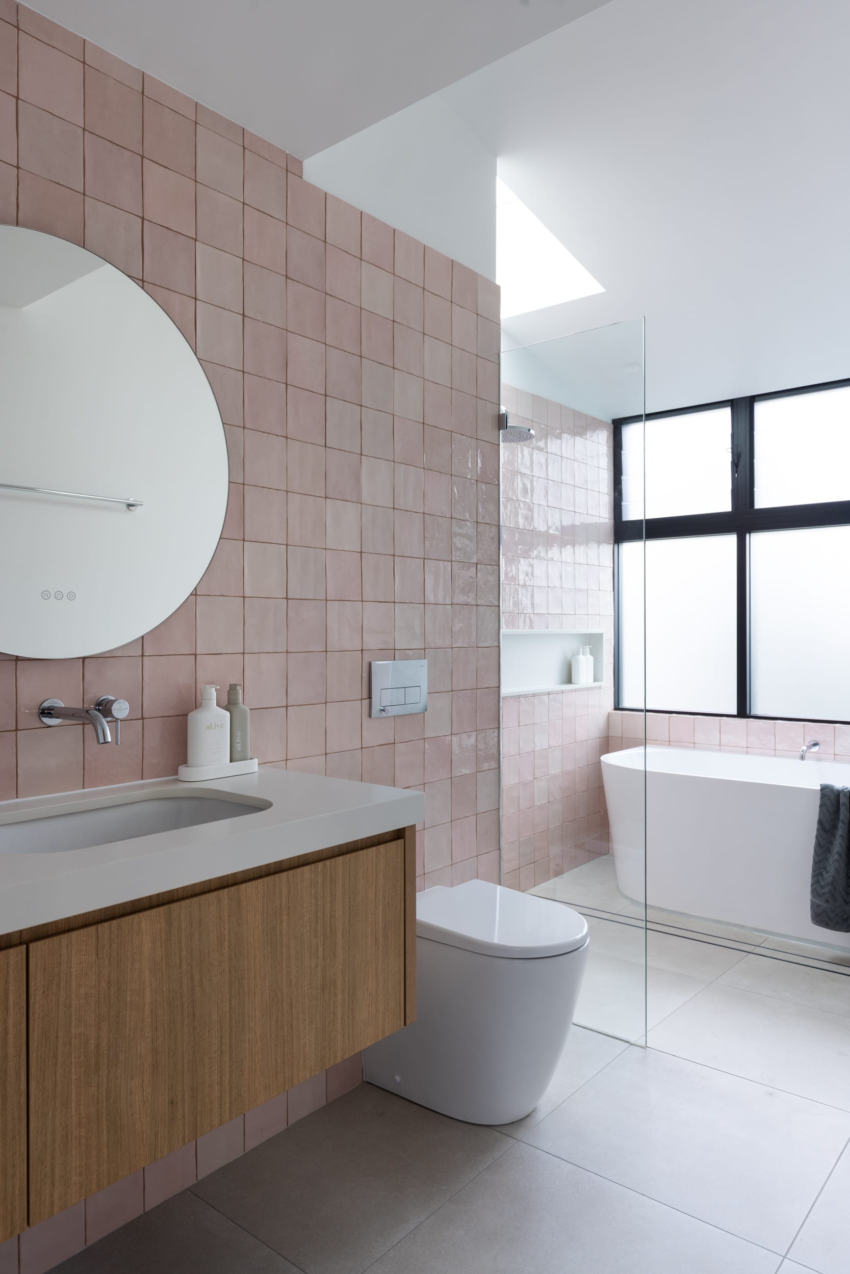 Bawley Point by Watershed Architects. Photography by Simon Whitbread. Bathroom with grey floor tiles, pink wall tiles, timber cabinetry, white countertop and toilet alongside glass screen. 