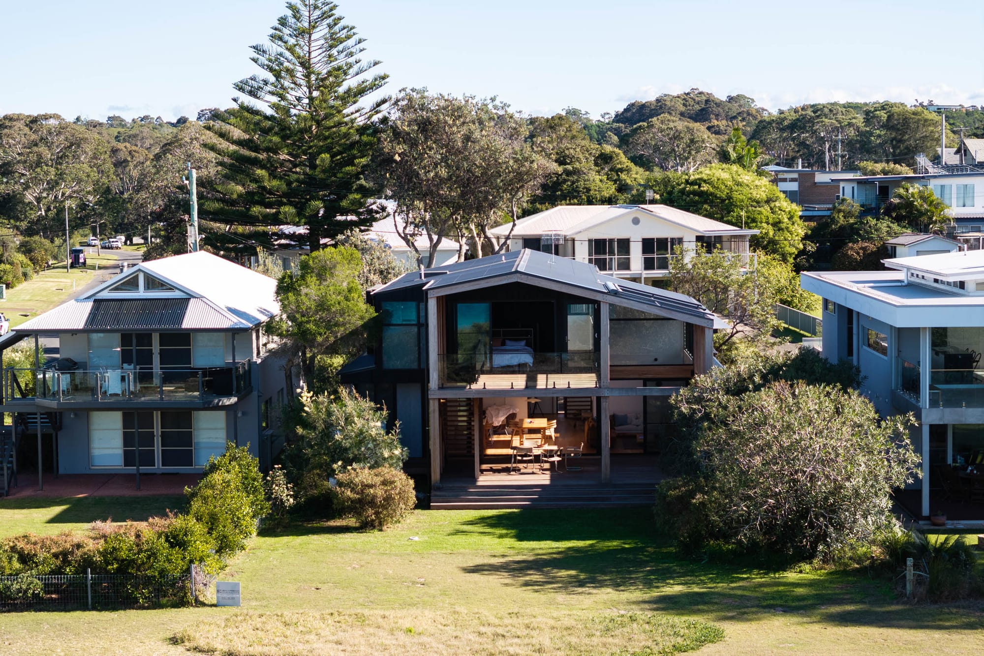 Bawley Point by Watershed Architects. Photography by Simon Whitbread. Aerial view of contemporary double-storey home with large balcony and timber deck, shouldered by double-storey homes on either side and facing onto grassed foreshore. 