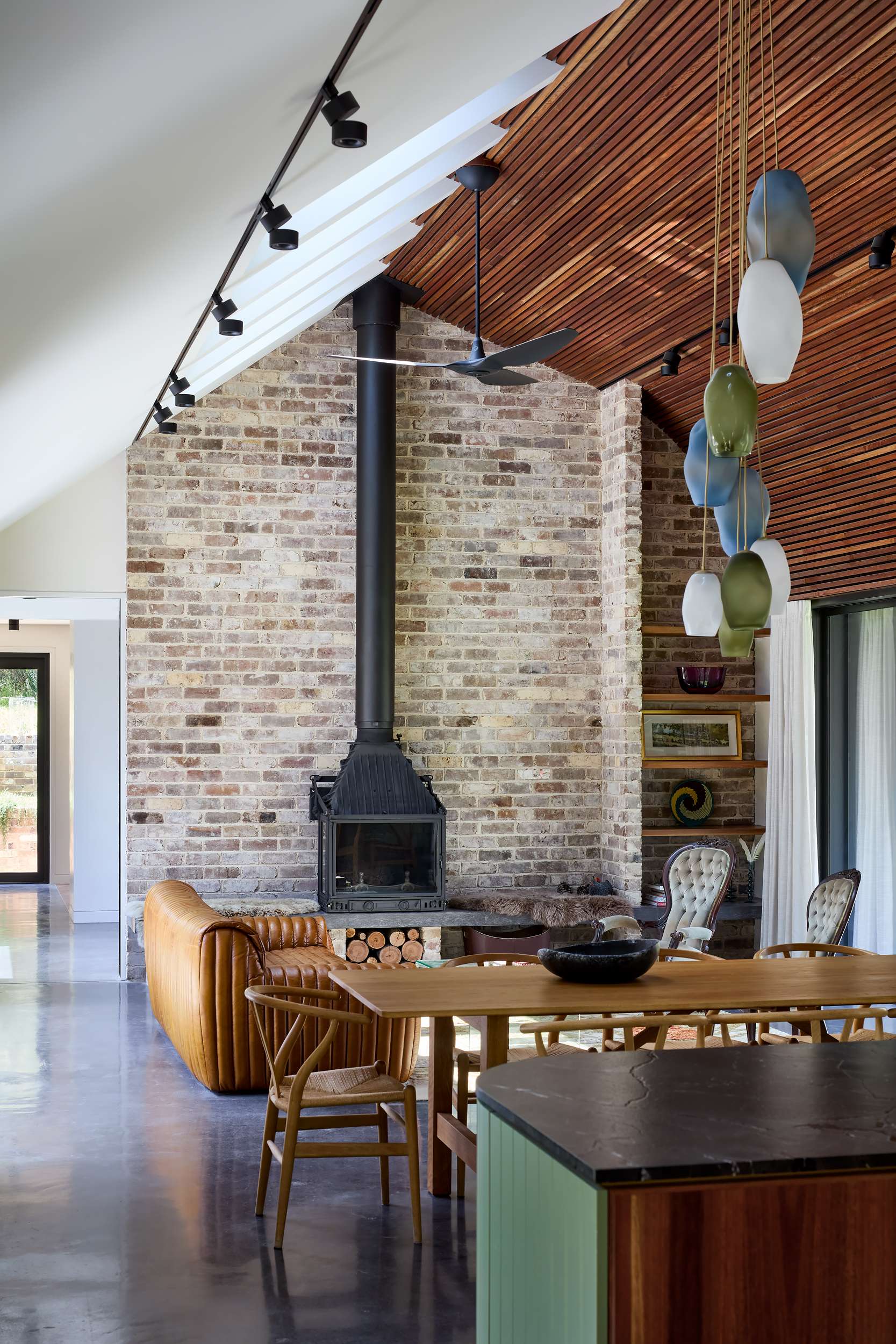 House Kimberley by AO Design Studio. Photography by Luc Remond. Open plan kitchen, dining and living area with polished concrete floors, timber dining table, brown leather couch, brick wall and timber clad ceiling.