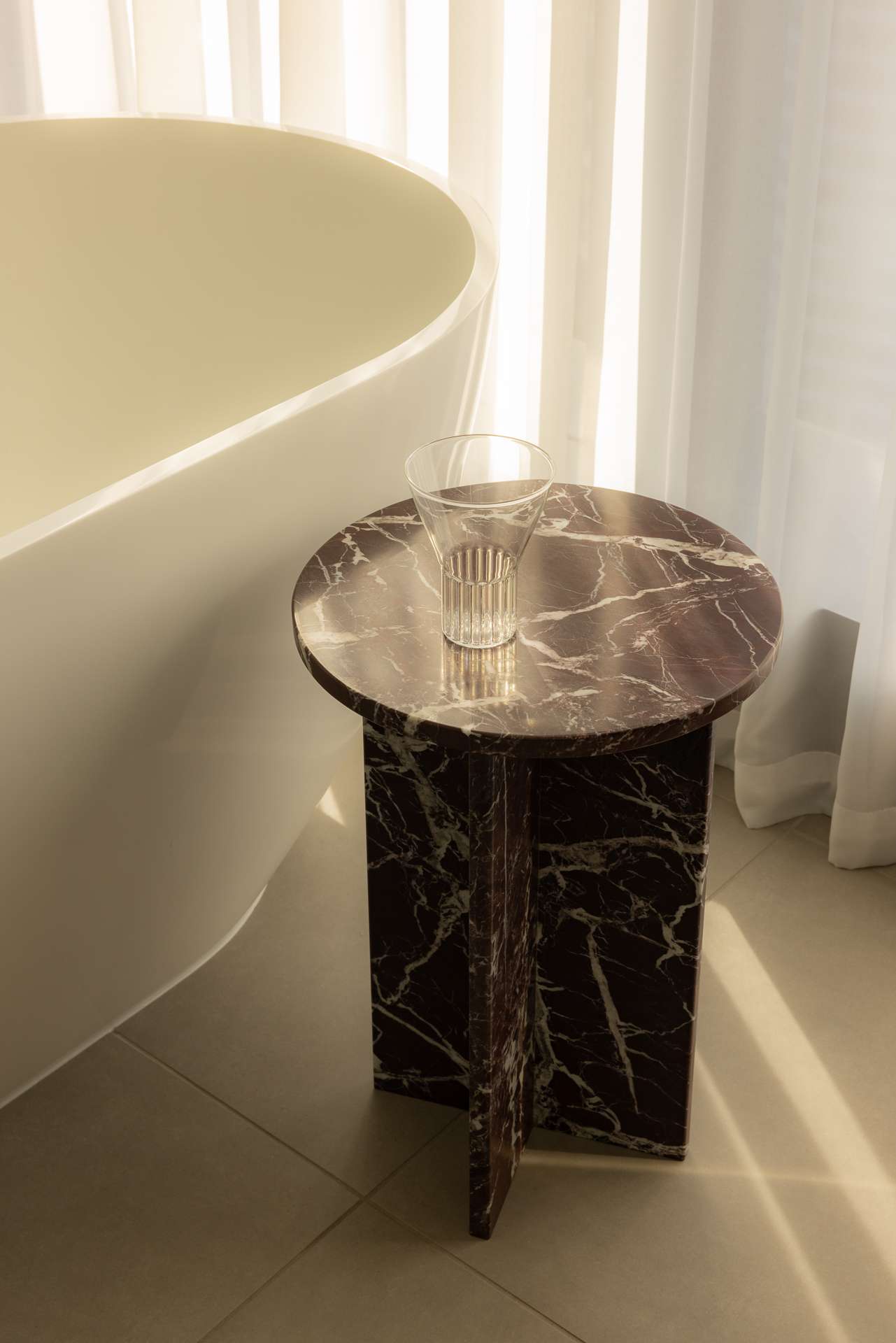 Cloud Residence by FURNISHD. Photography by Dylan James. Dark brown round side table with thick, white veining, positioned alongside white tub and white sheers in bathroom with white tiled floors.