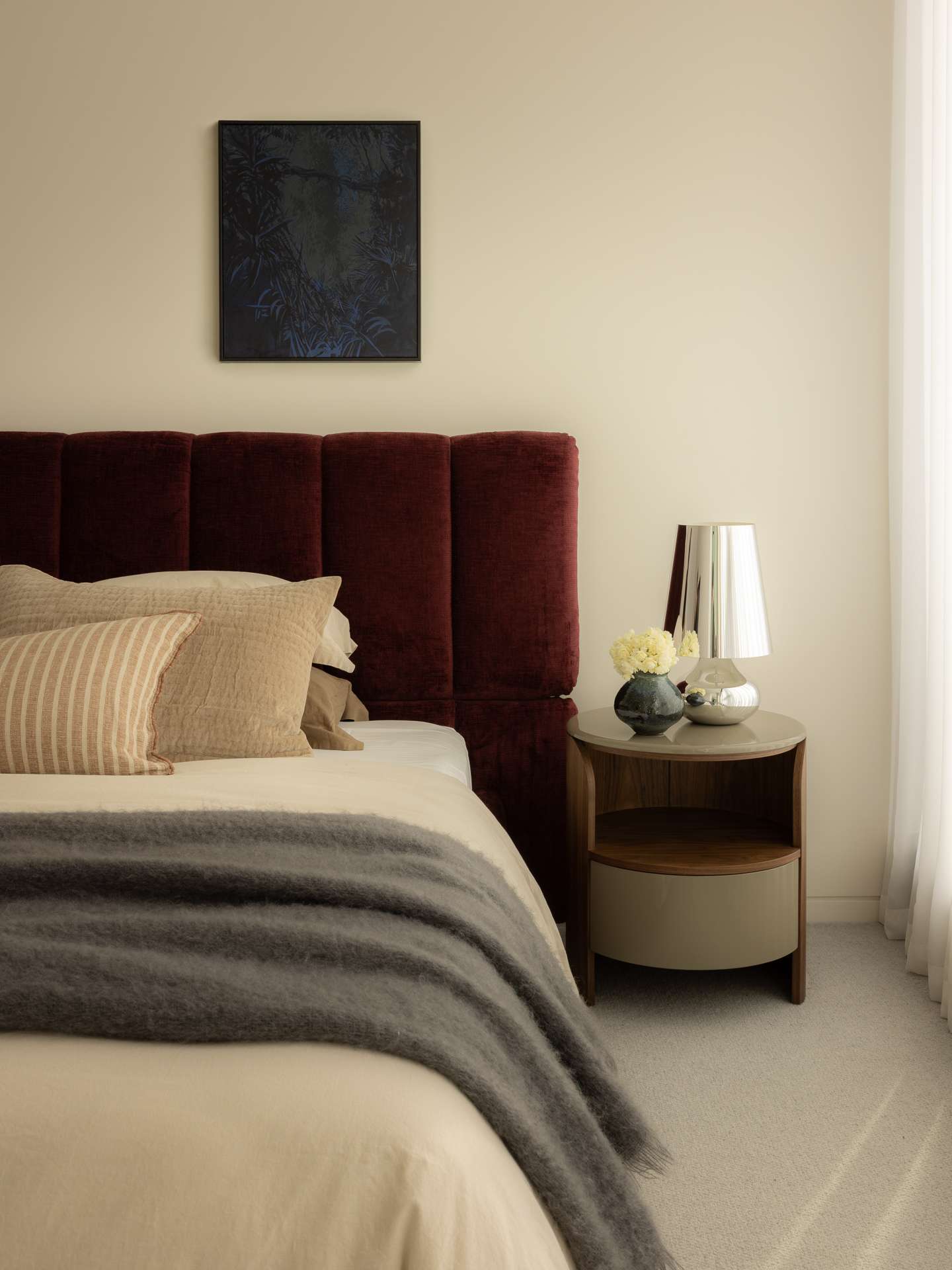 Cloud Residence by FURNISHD. Photography by Dylan James. Dark red upholstered bedframe against white wall, and a round timber side table with a chrome lamp.