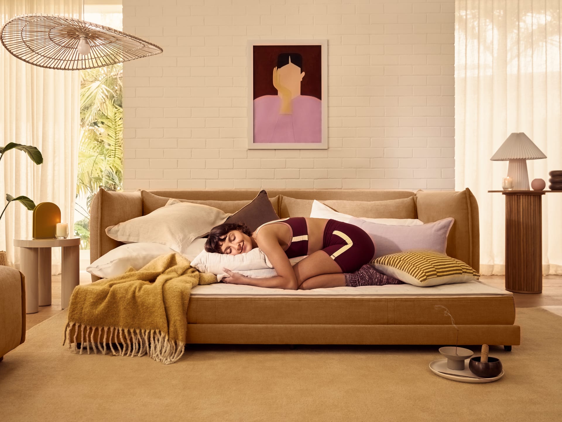 Koala Byron Sofa Bed [3rd Gen] in Bronzed Aussie. Woman in matching burgundy set lying crouched on her stomach on mustard couch with beige cushions in front of white brick wall. 