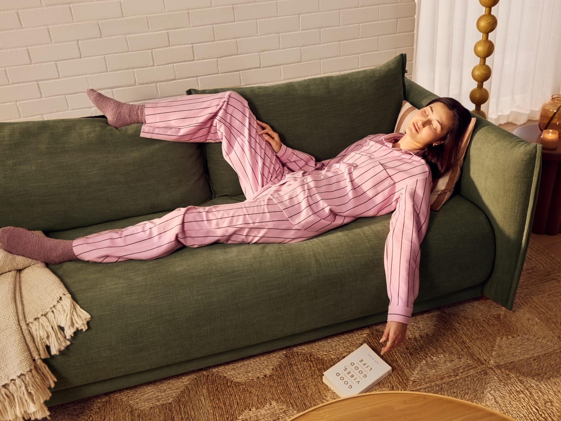 Koala Byron Sofa Bed [3rd Gen] in Forest Walk. Woman in pink stripey pyjama set asleep on dark olive couch, in room with white brick wall and patterned jute rug. 