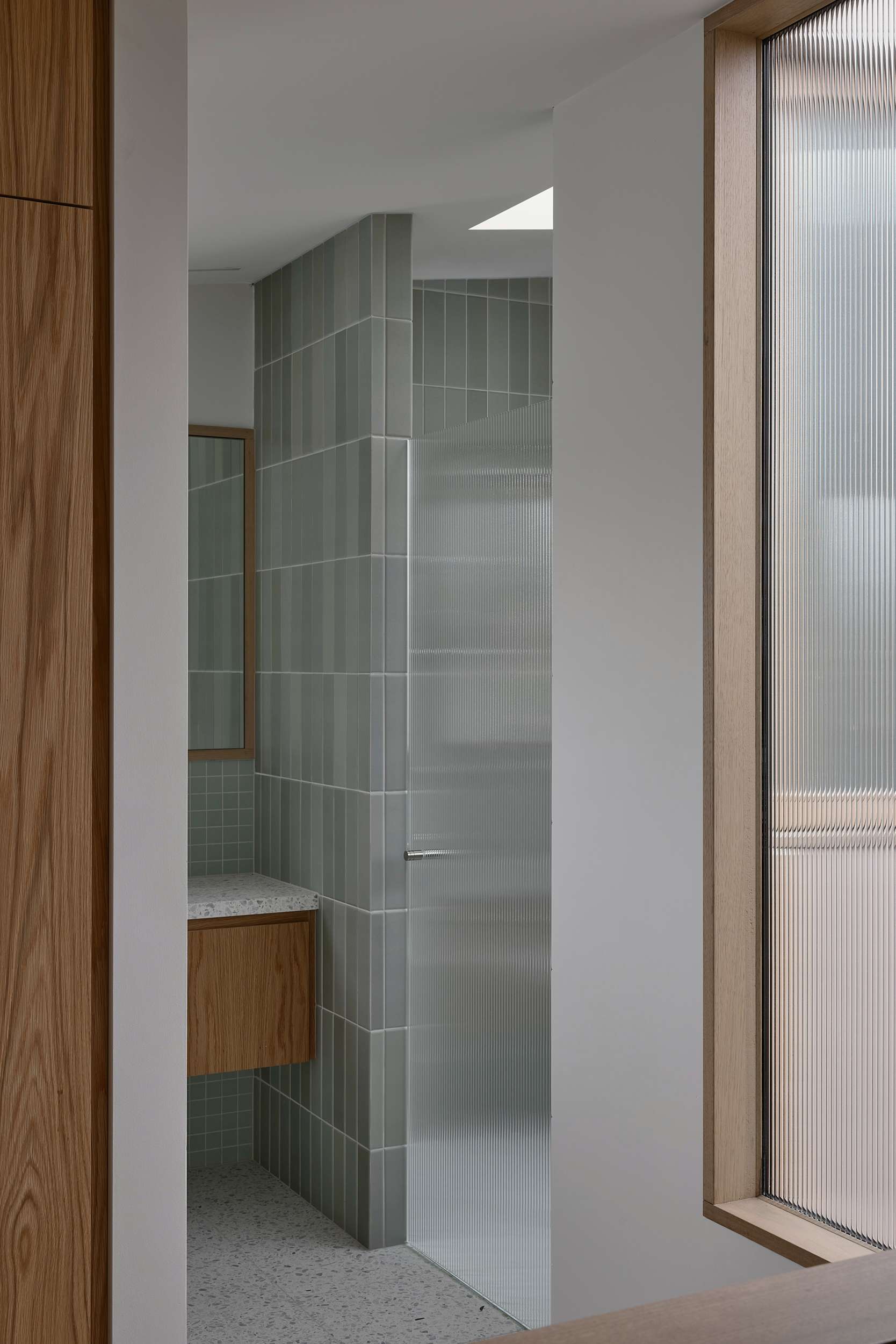 Home Pavilion by MRTN Architects. Fluted glass door in bathroom with white terrazzo flooring, timber cabinets, white countertops and green wall tiles. 