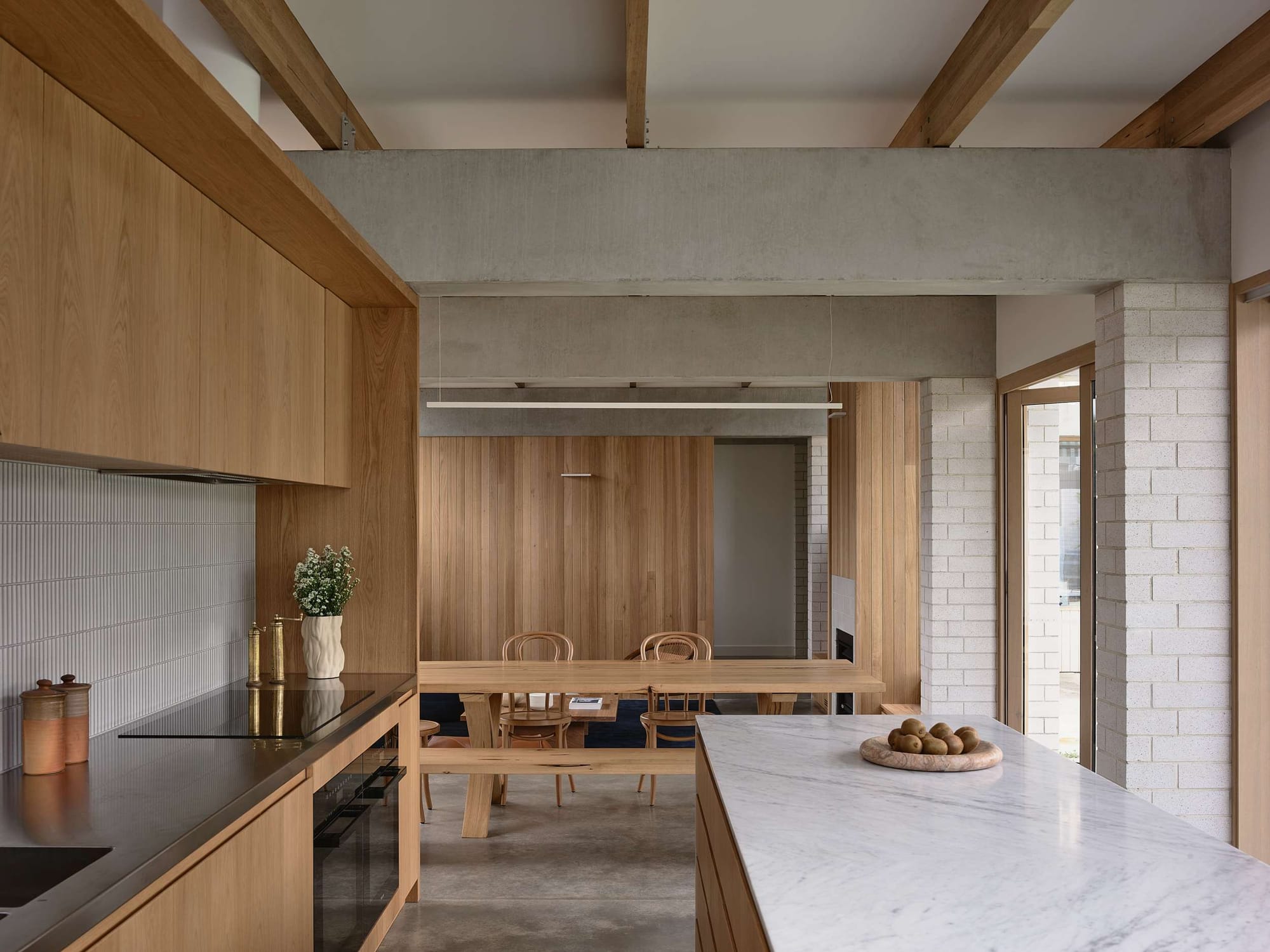 Home Pavilion by MRTN Architects. View across kitchen with timber cabinetry, white stone countertops and finger-tile splashback, across to dining space with timber dining table, exposed concrete beams and brick pillars. 