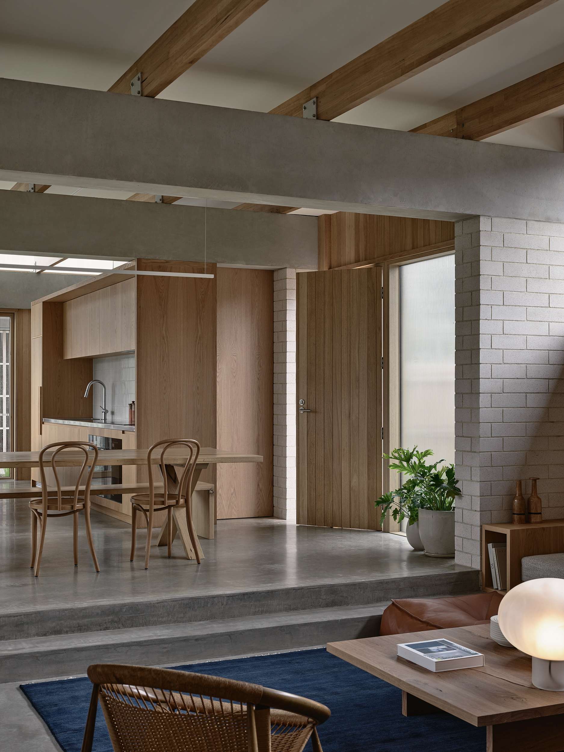 Home Pavilion by MRTN Architects. View across sunken living space with concrete floors, navy rug and timber furnishings up to elevated dining and kitchen with exposed concrete beams and timber details. 