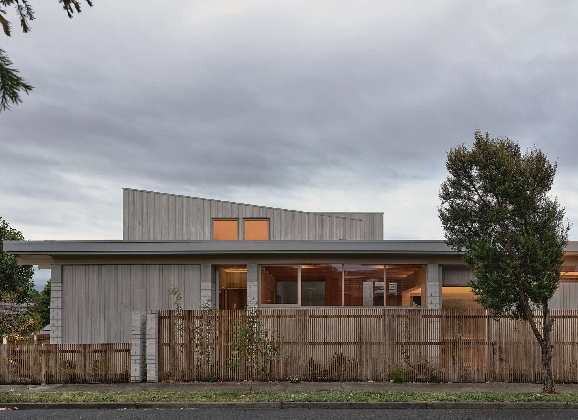 Home Pavilion by MRTN Architects. Front facade of contemporary home with brick, timber and concrete facade, and an asymmetrical roofline, behind a timber fence. 