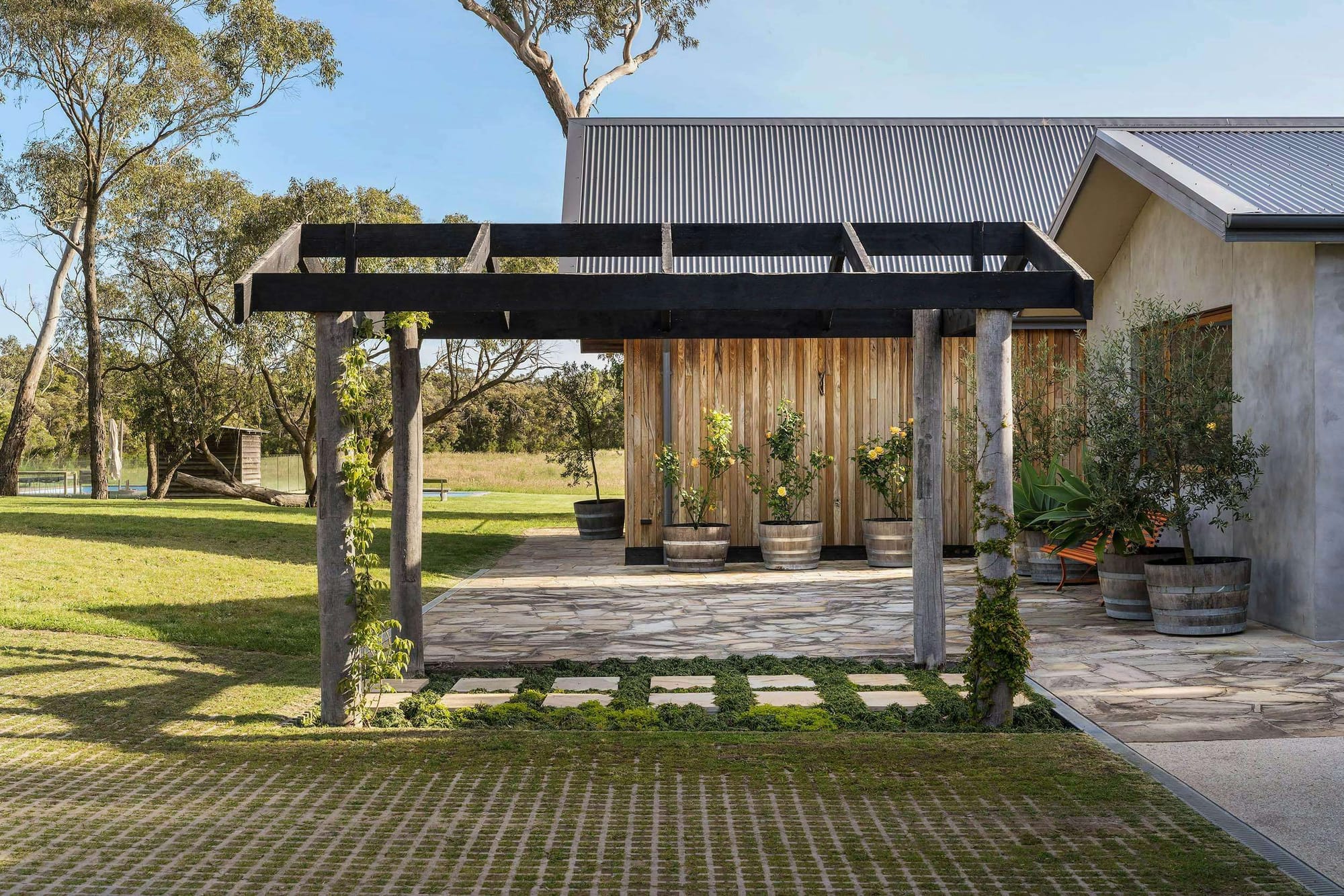 Mooroorduc Farm by Merrylees Architecture. Photography by Mitch Lyons. Timber pergola in front of paved patio area, extending from clay and timber clad structure in rural setting.