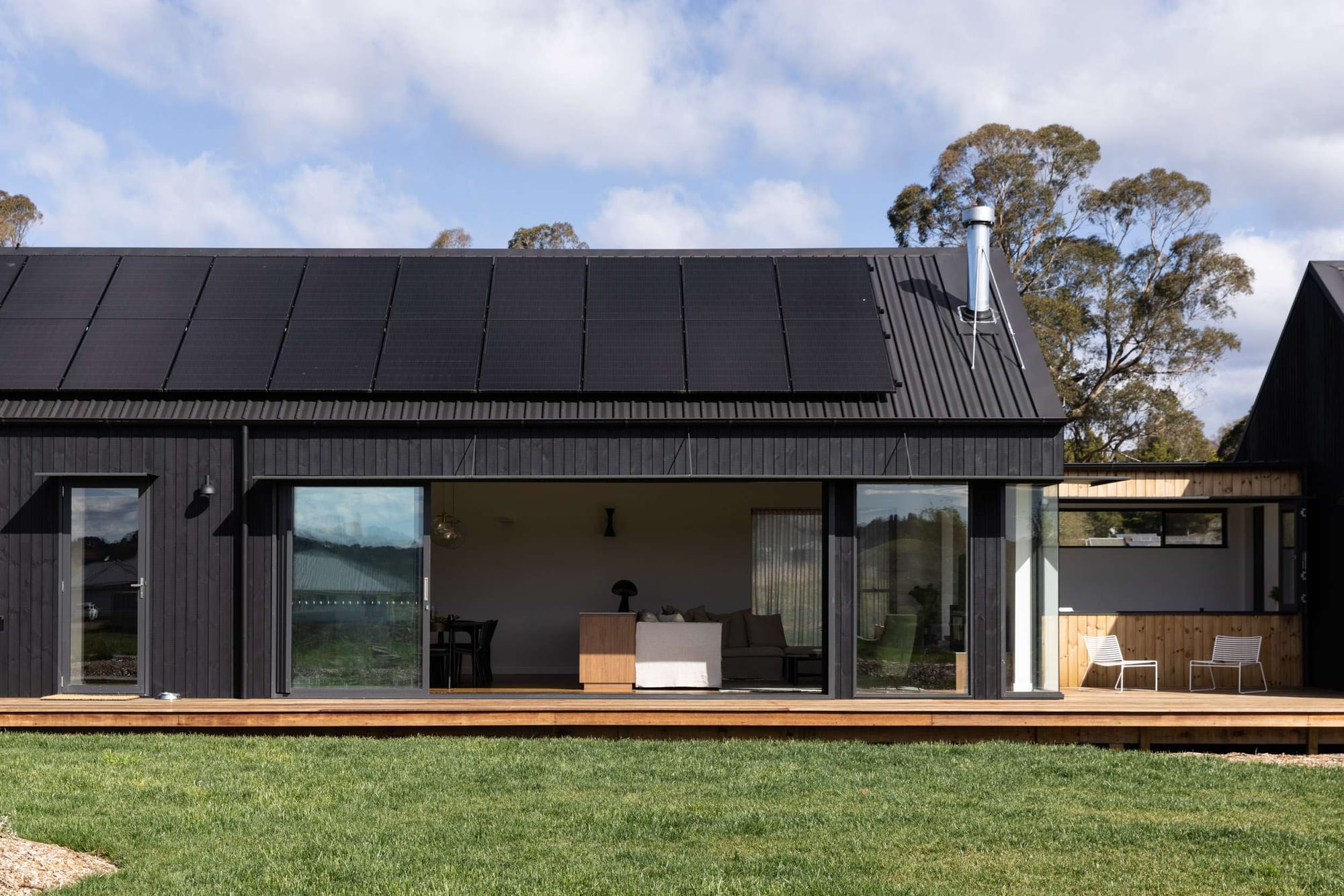 Birrigai House by Studio ERA. Photography by Simon Whitbread. Long, black pavilion style home with simple pitched black roof, timber clad wall behind entrance, and grassed garden.