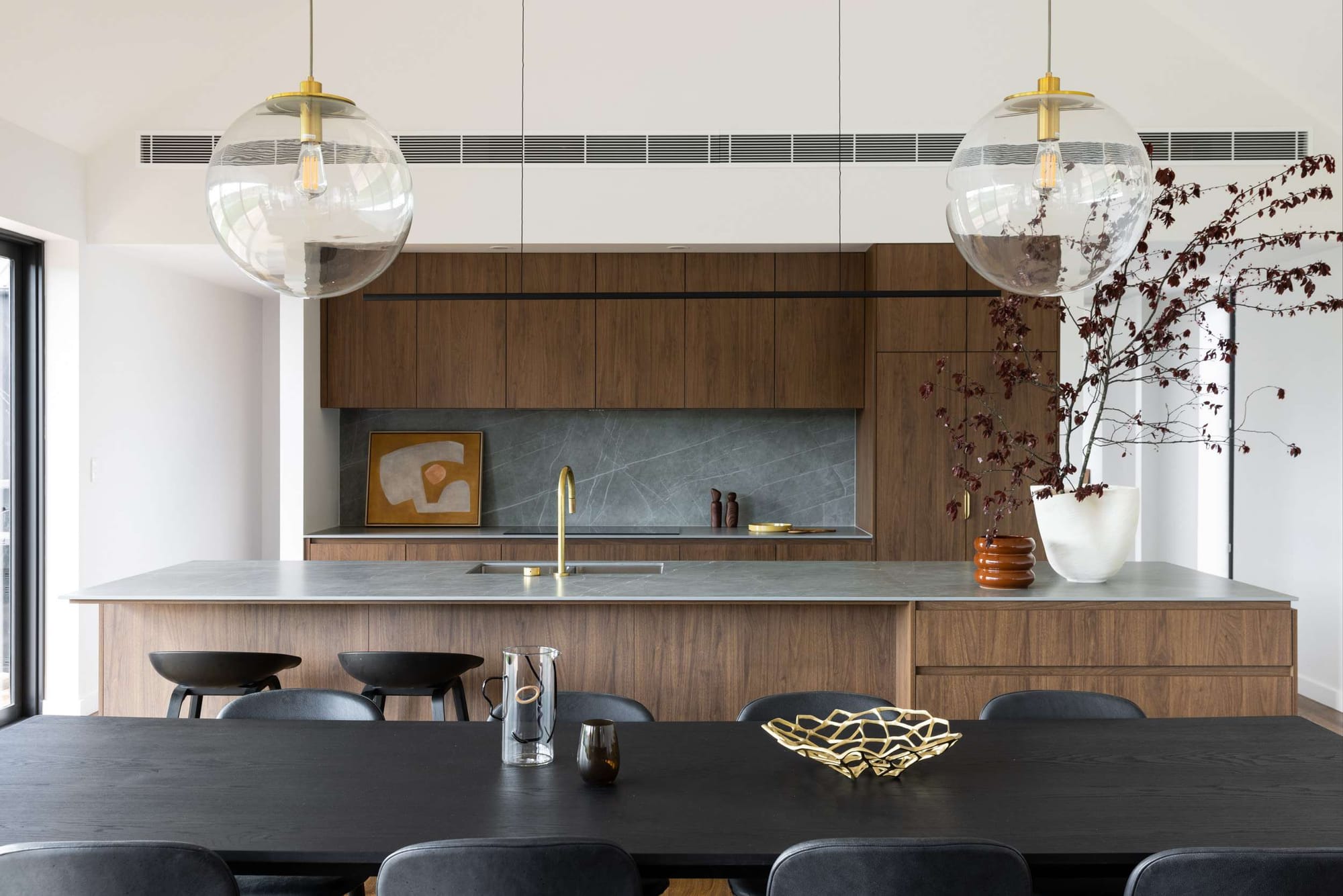 Birrigai House by Studio ERA. Photography by Simon Whitbread. Kitchen with timber cabinetry, grey stone countertop and splashback, two glass spherical pendants, and black dining table.