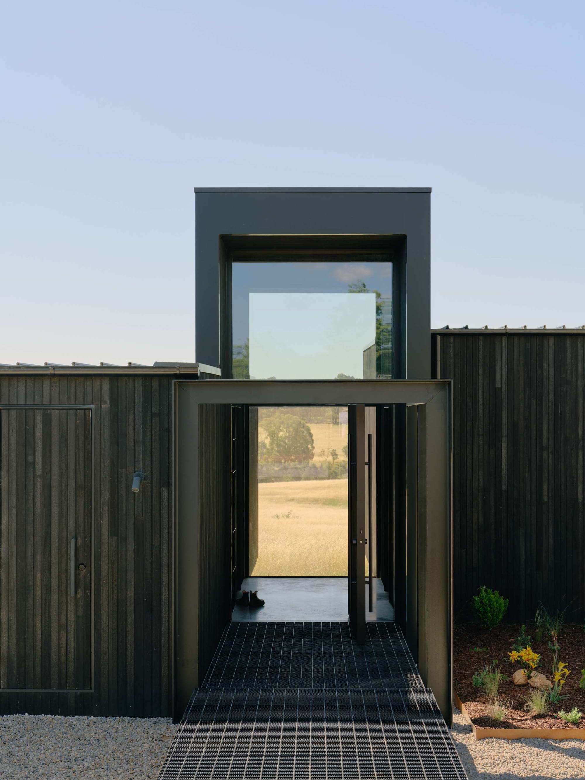 Marraweeney by Tom Robertson Architecture. Photography by Tom Ross. Exterior facade of home and entrance, with tall black archway and walkway leading into pavilion home with black timber exterior.
