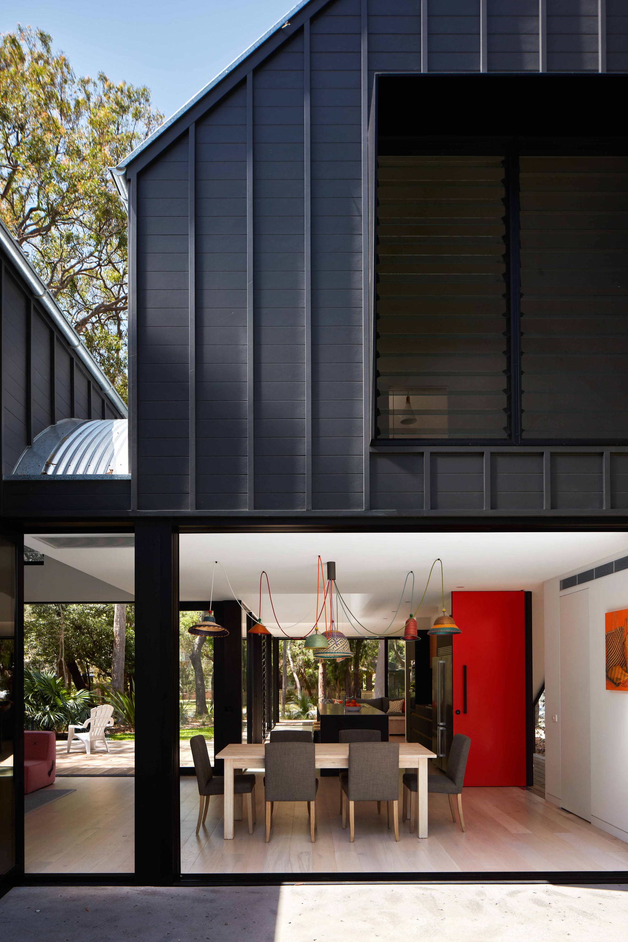 Pearl Beach II by COCO Architecture. Dinning are with large windows. Wooden dinning table with grey chairs. Red door and colourful lamps.