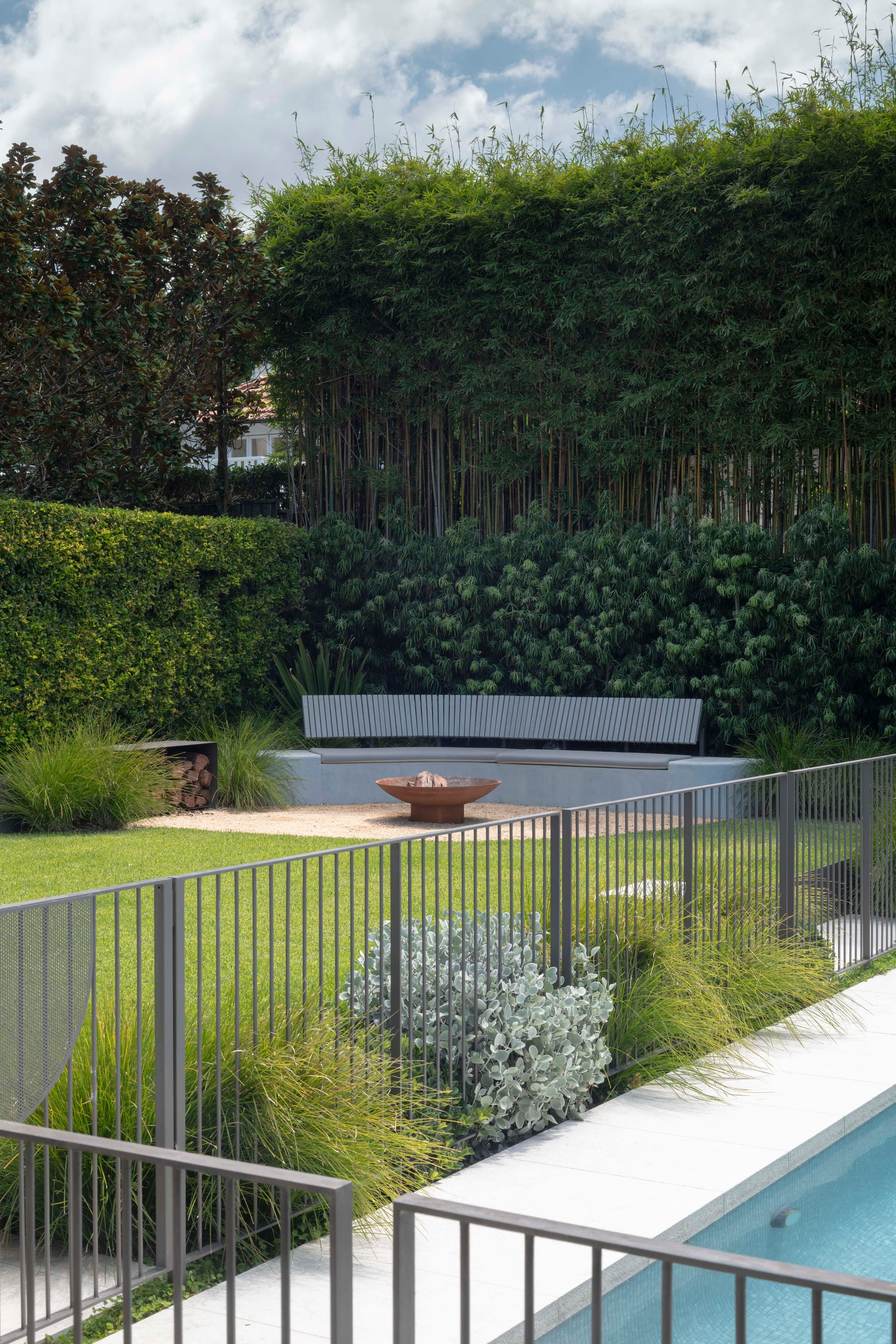 Fire pit with logs and grey sitting area. Pool with grey fencing and bushes. Green grass and hedges.