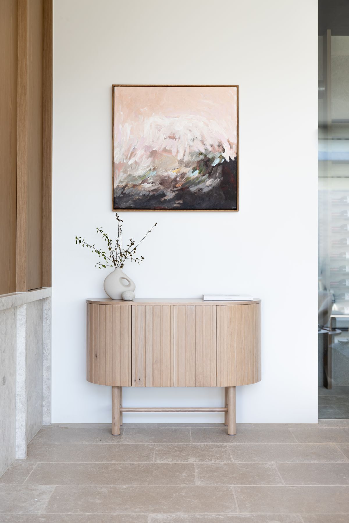 wooden sideboard against a wall. Above the sideboard hangs a framed abstract painting with hues of pink, white, and gray.